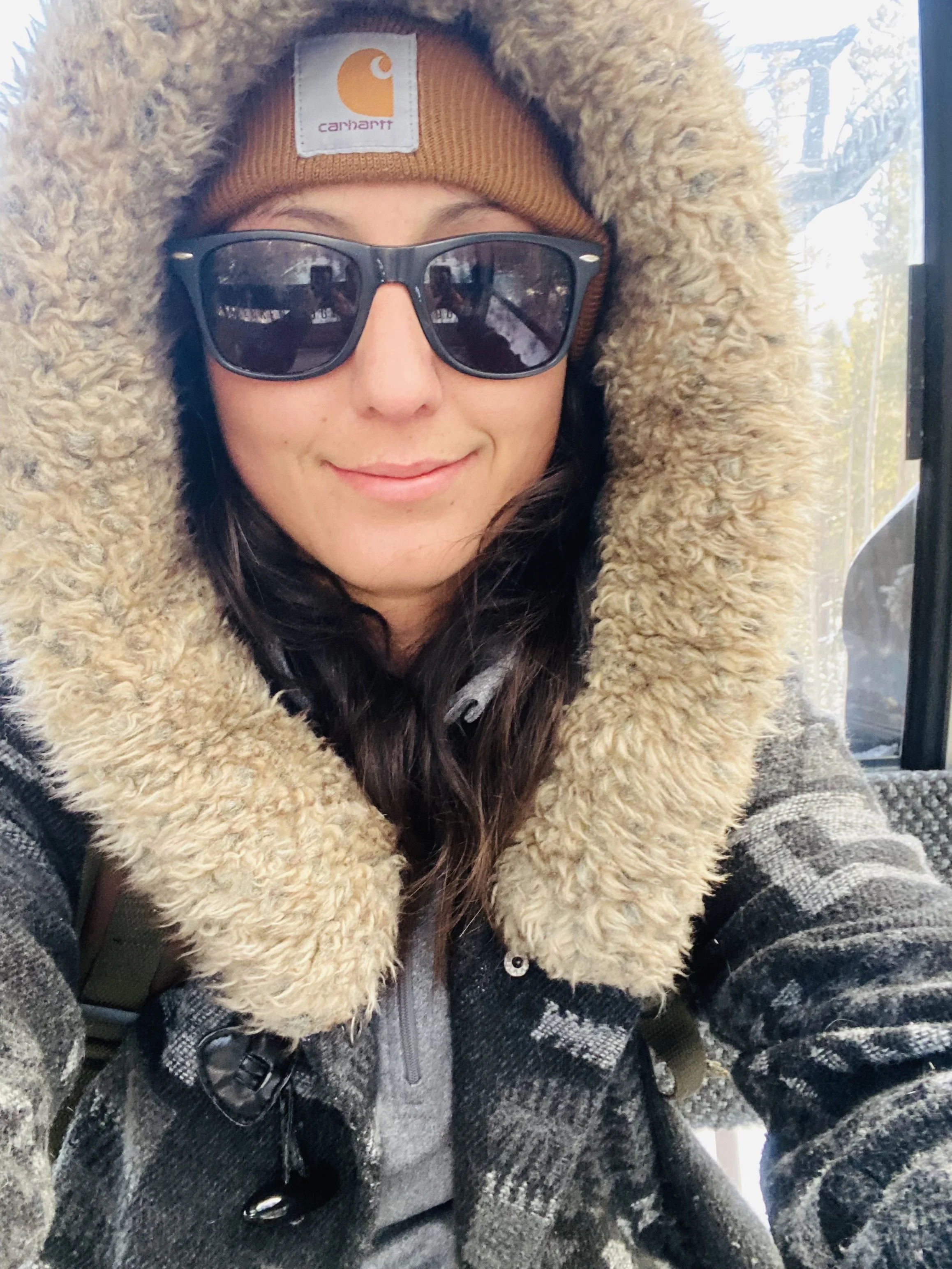 A woman dressed in winter attire, including a brown Carhartt beanie, black sunglasses, and a coat with a large fur-lined hood, sitting inside a bus or train with a window showing a winter scene outside.