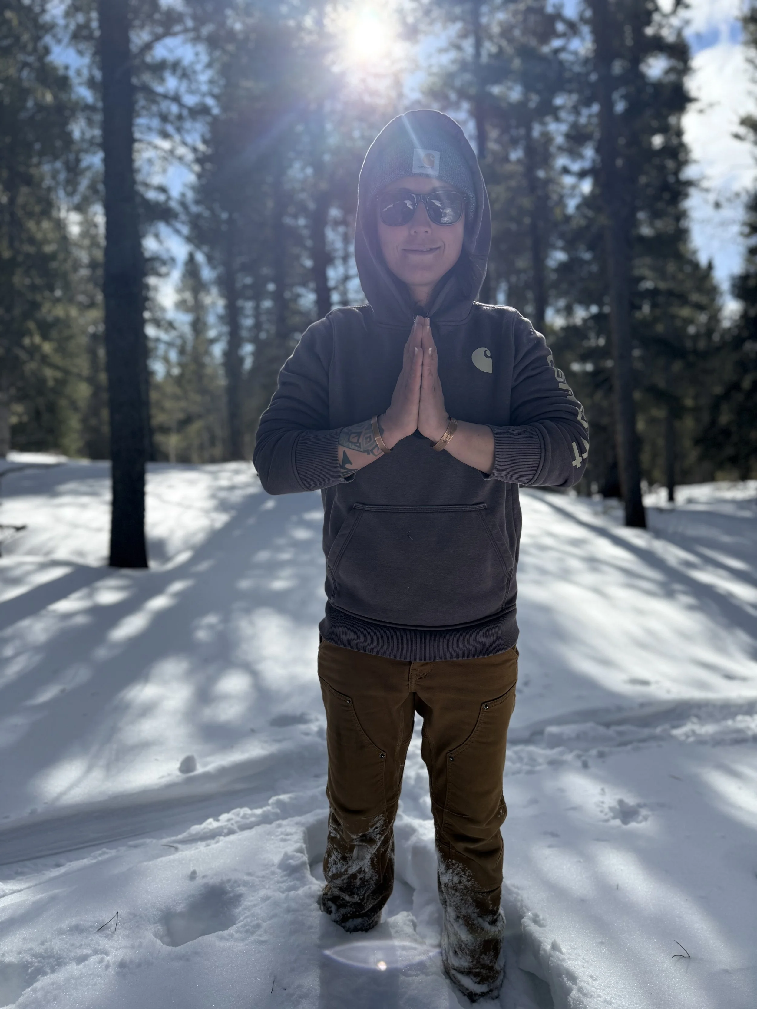 Person standing in snowy forest with hands in prayer position, wearing sunglasses, dark hoodie, and brown pants, with sunlight shining through trees behind.