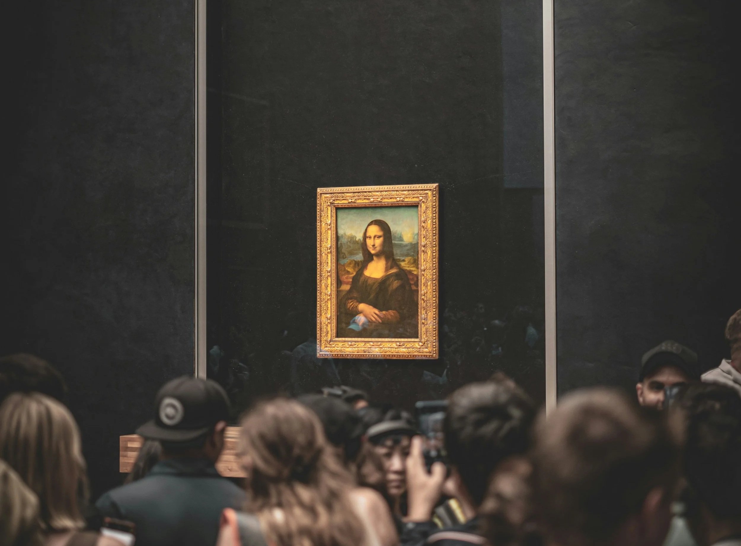 The image shows the Mona Lisa painting displayed in a darkened exhibition room of an art museum, surrounded by a crowd of visitors watching it.