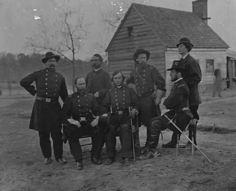 Black and white photo of seven men in military or period clothing, some seated and some standing, in front of a wooden house with leafless trees in the background.