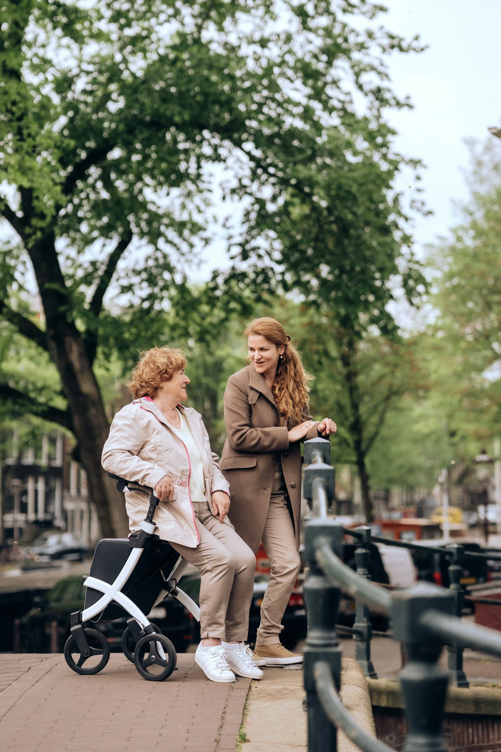 Two women, one elderly and one middle-aged, are standing on a sidewalk by a canal, engaging in conversation with a background of green trees and canal boats.
