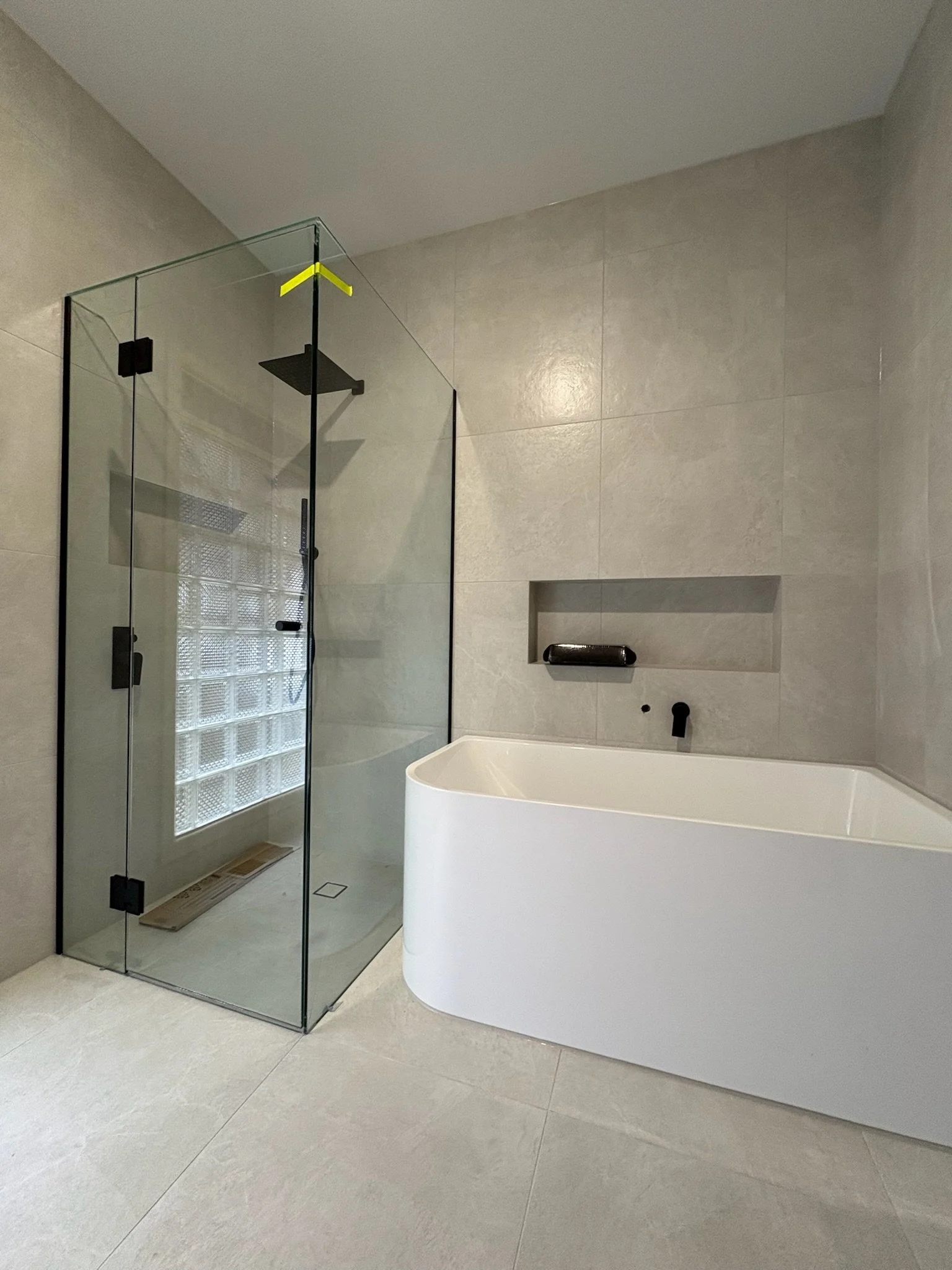 Modern bathroom with glass-enclosed shower and white bathtub with curved edge, beige tiled walls, and built-in niche. All joinery and tiled areas have been caulked and sealed by a professional caulker.