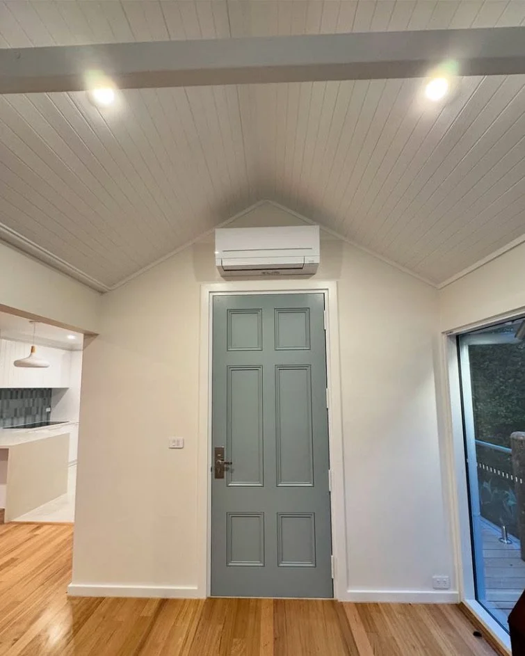 Interior view of a room with a gray front door, white walls, wooden floor, a split air conditioner above the door, and a sliding glass door on the right leading to an outside balcony.
