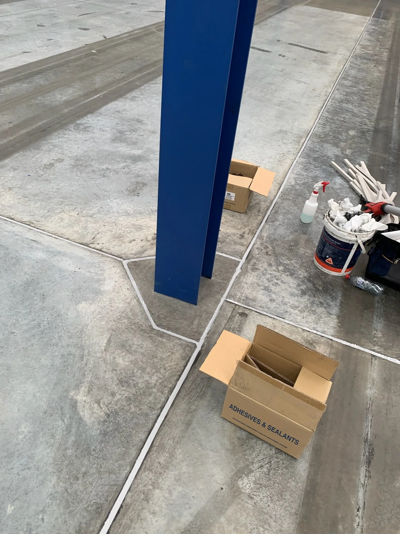 Construction area with a blue metal post, a cardboard box labeled 'Adhesives & Sealants', and various tools and supplies. Wide concrete joints have been sealed by a professional caulker.