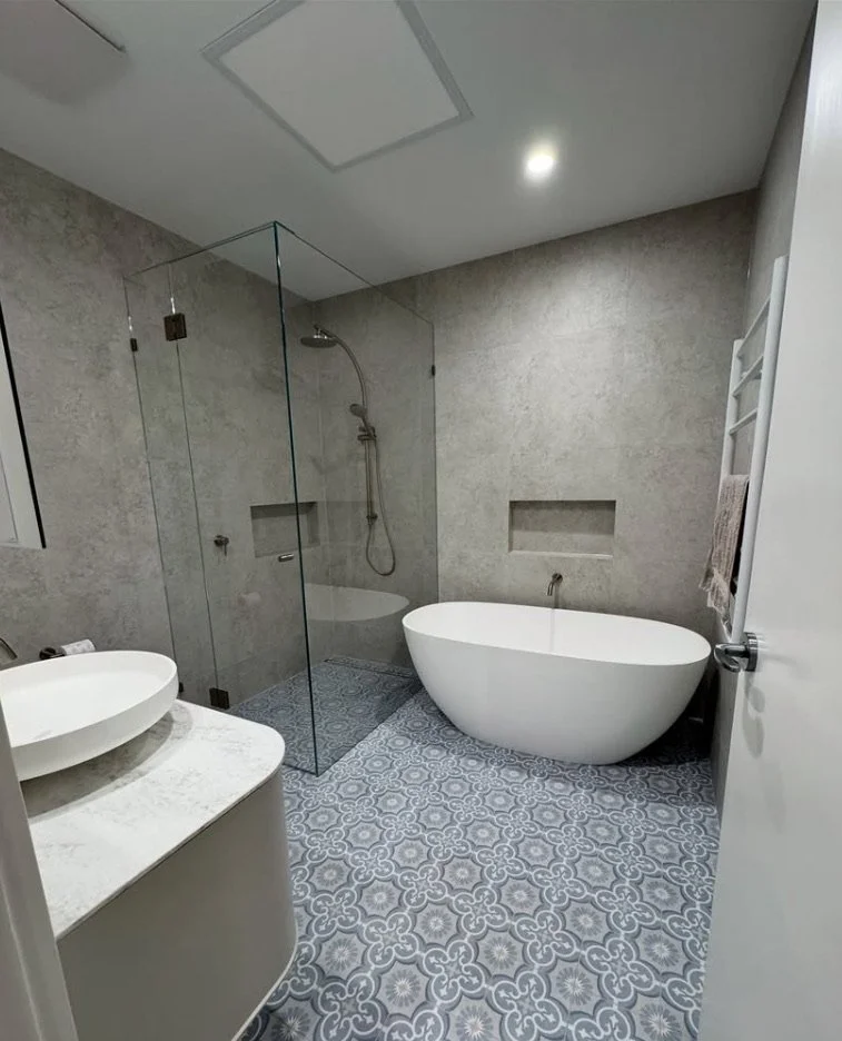 Modern Bayside bathroom with a glass-enclosed shower, a free-standing bathtub, sink on a marble countertop, and light gray tiled walls and patterned floor tiles. All joints and tiled areas have been caulked and sealed by a professional caulker
