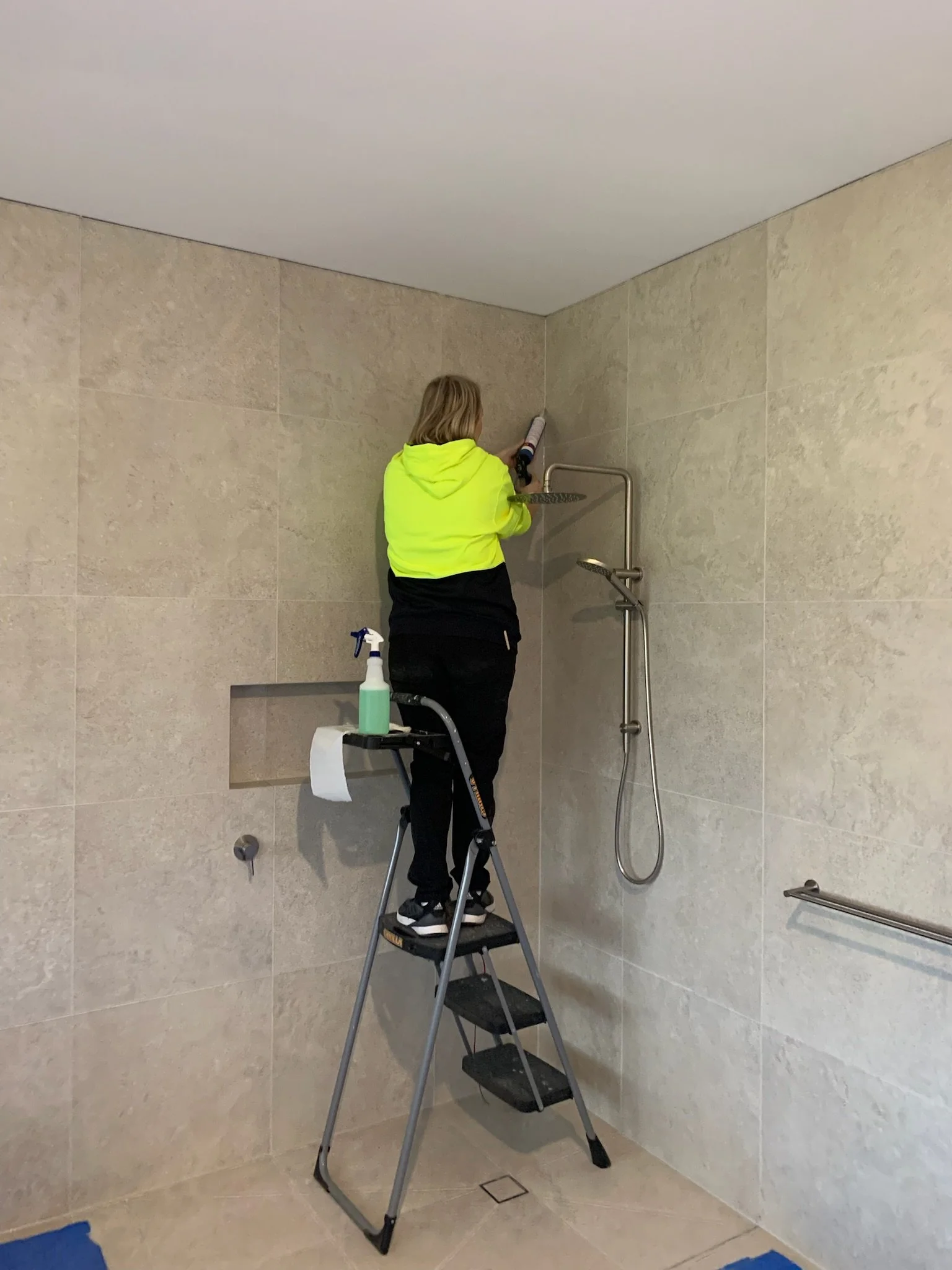 A woman standing on a step ladder caulking the corner of a shower with a caulking gun. She is wearing a bright yellow hoodie and black pants.