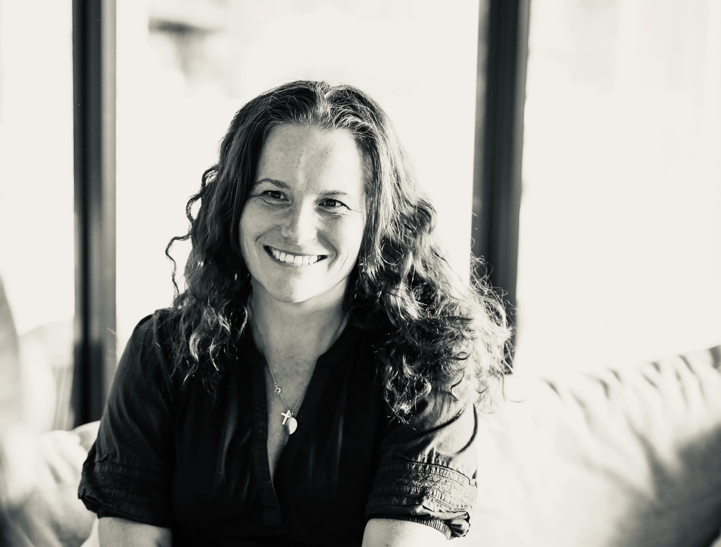 A smiling woman with long curly hair wearing a dark jacket and a cross necklace inside a bright room with windows and some furniture.