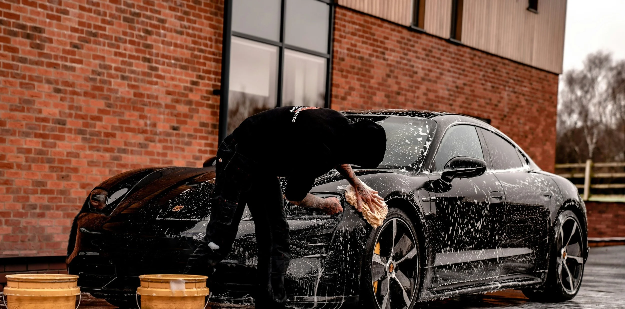 Person washing a black sports car with soap and a sponge outside. There are two yellow buckets in front of the car, and a brick building in the background.