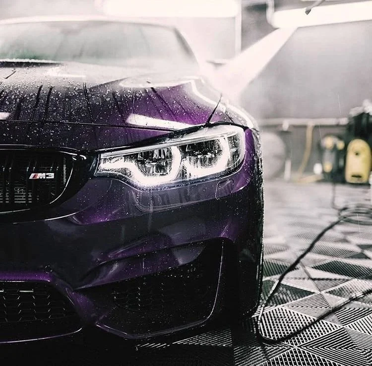 A purple BMW M3 car being washed, with water droplets on its surface and a spray of water in the background.