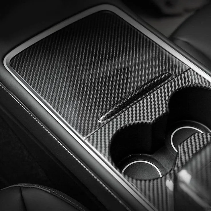 Close-up of a car's center console with a carbon fiber finish, featuring a sliding cover and two cup holders.