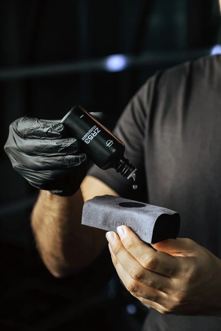A person wearing black gloves applies a liquid from a small bottle onto a foam applicator pad.
