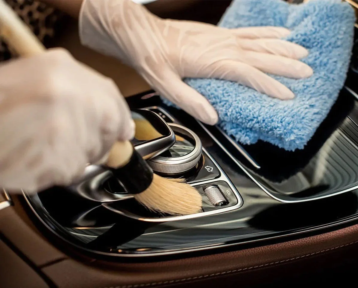 Person wearing white gloves cleaning the center console of a car with a microfiber cloth and brushes.