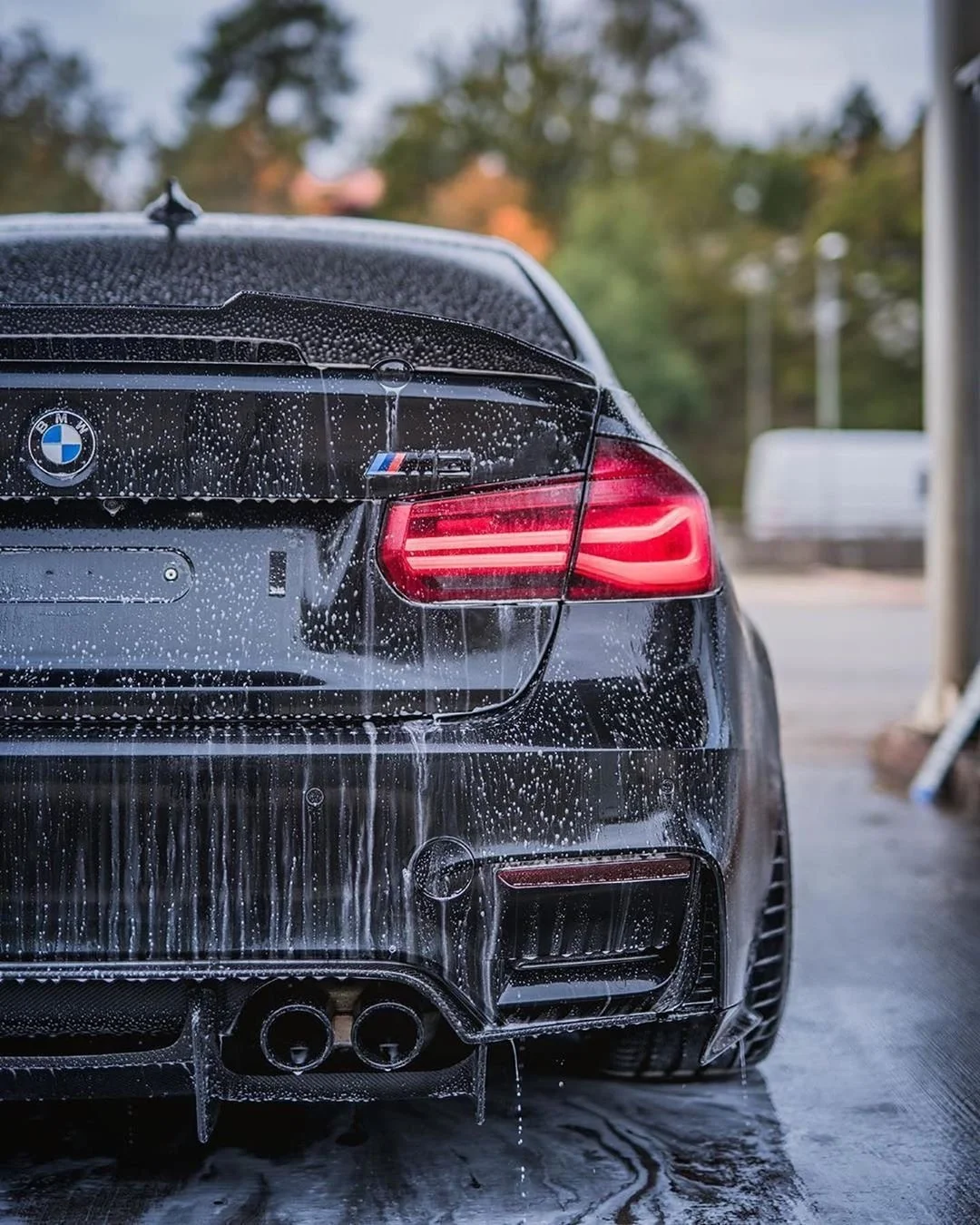 The rear end of a black BMW M3 sports car covered in soap suds during a car wash. The car's taillights are illuminated and water is dripping from the soap.