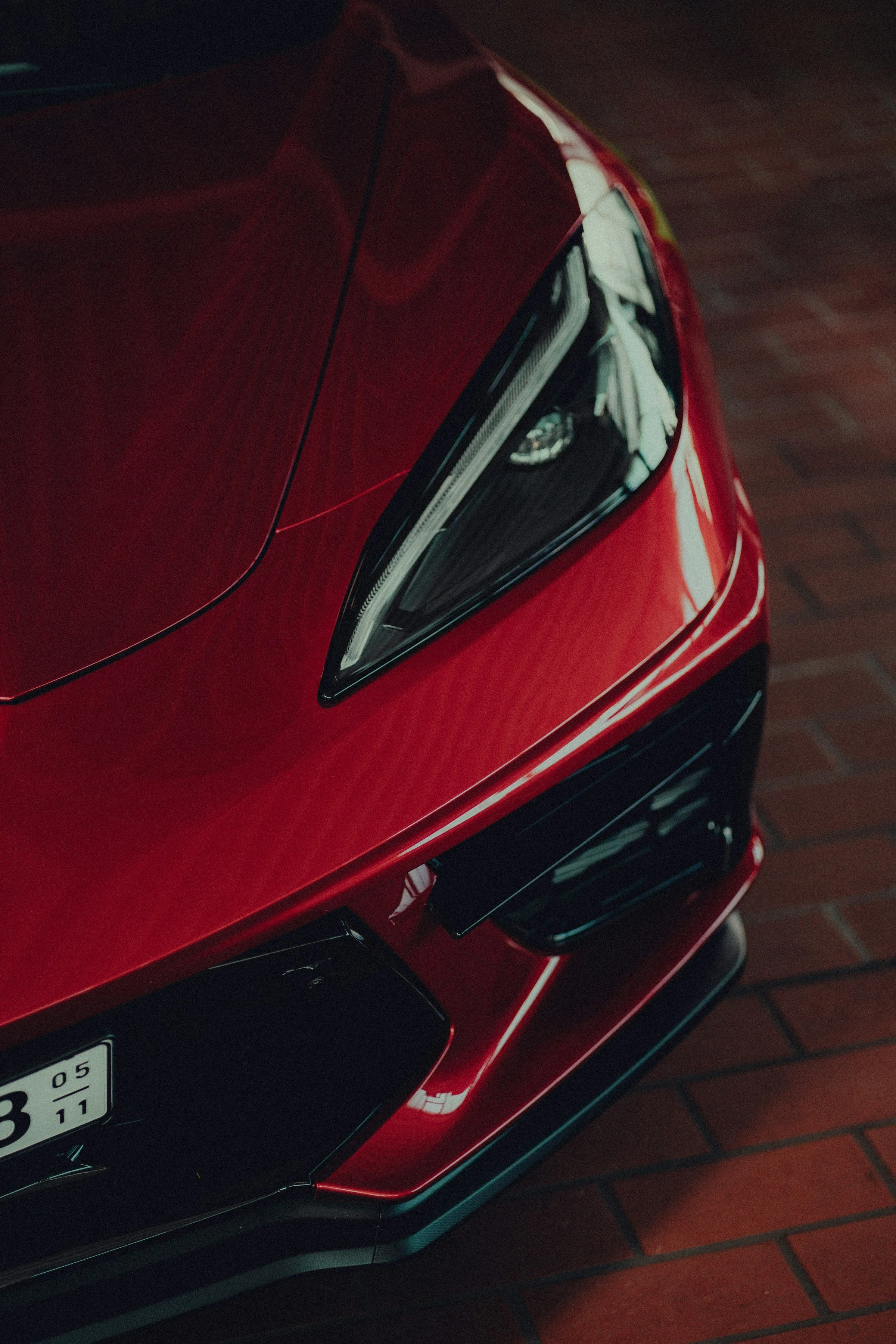 Close-up of the front corner of a red luxury sports car, showing part of the hood, headlight, and front bumper with reflections on its shiny surface, parked on a brick floor.
