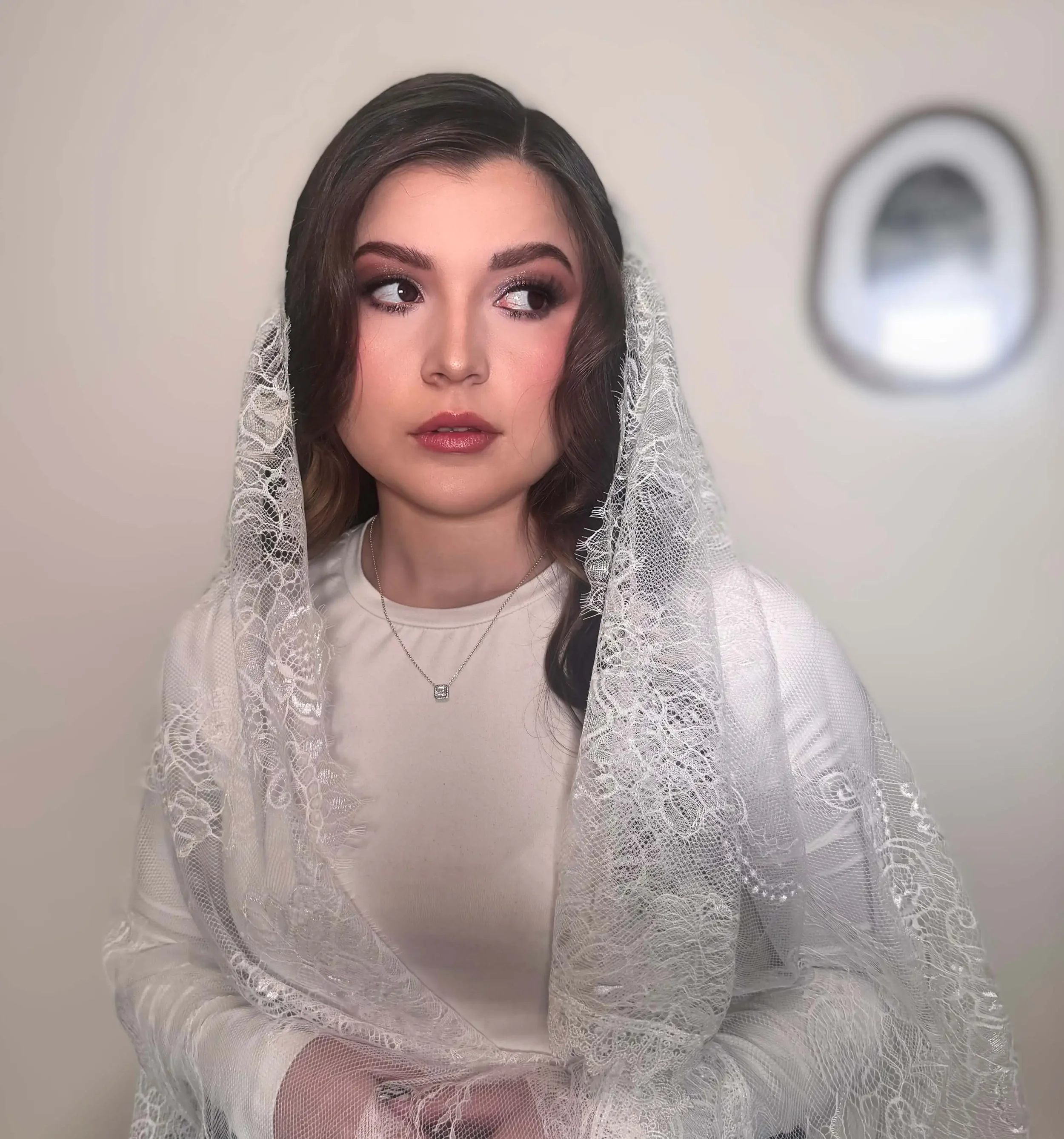 A woman wearing soft bridal makeup with smoky brown eyes, rosy nude lips, and glowing skin, framed by a delicate lace veil for a romantic, elegant look
