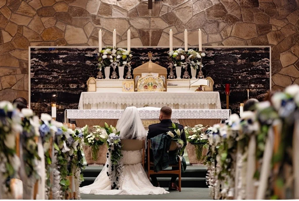 Bride in lace wedding gown and groom in traditional kilt seated together during a church wedding ceremony.