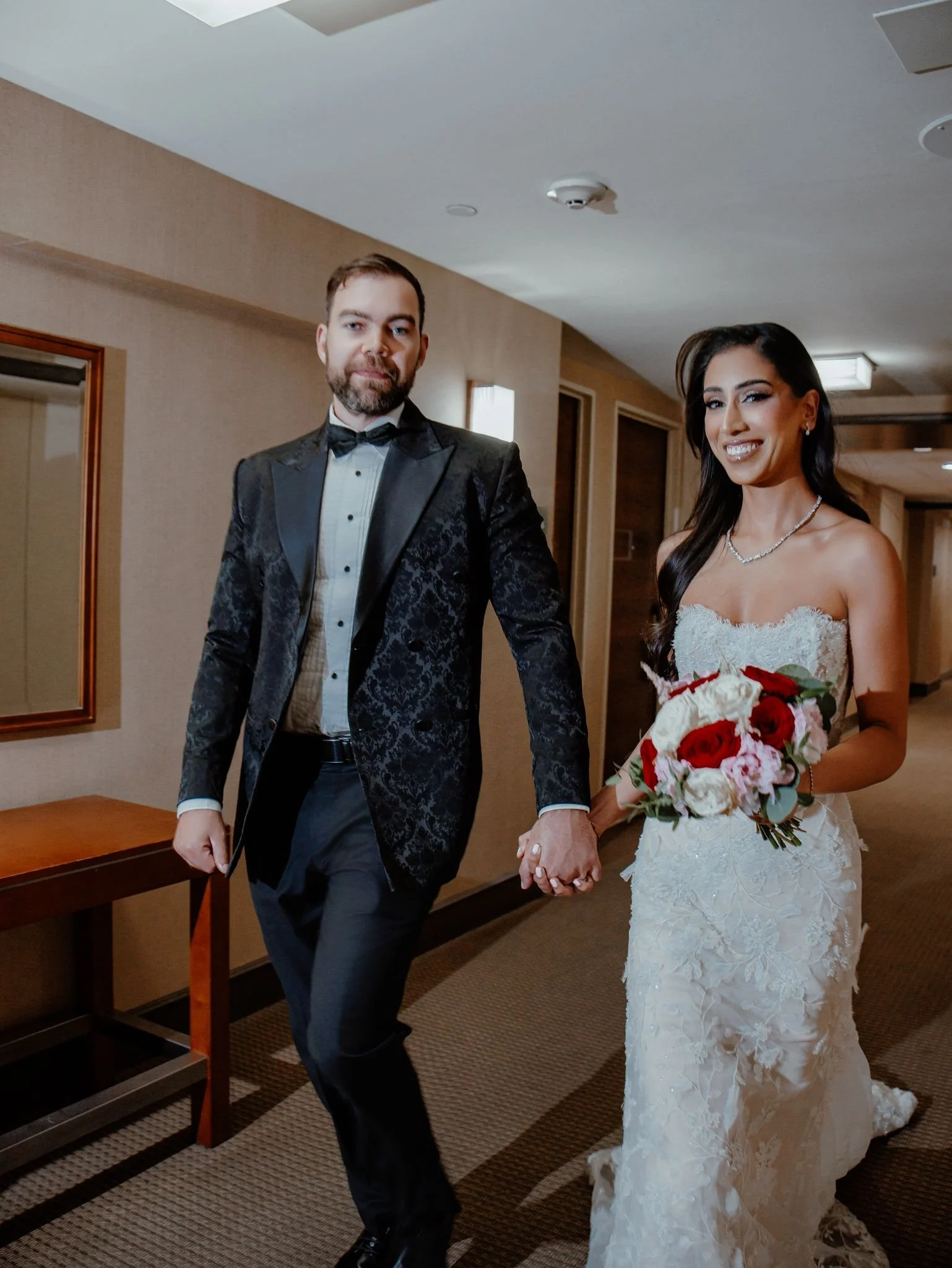 Bride in lace wedding gown and groom in formal tuxedo walking hand i.n hand down a hotel hallway