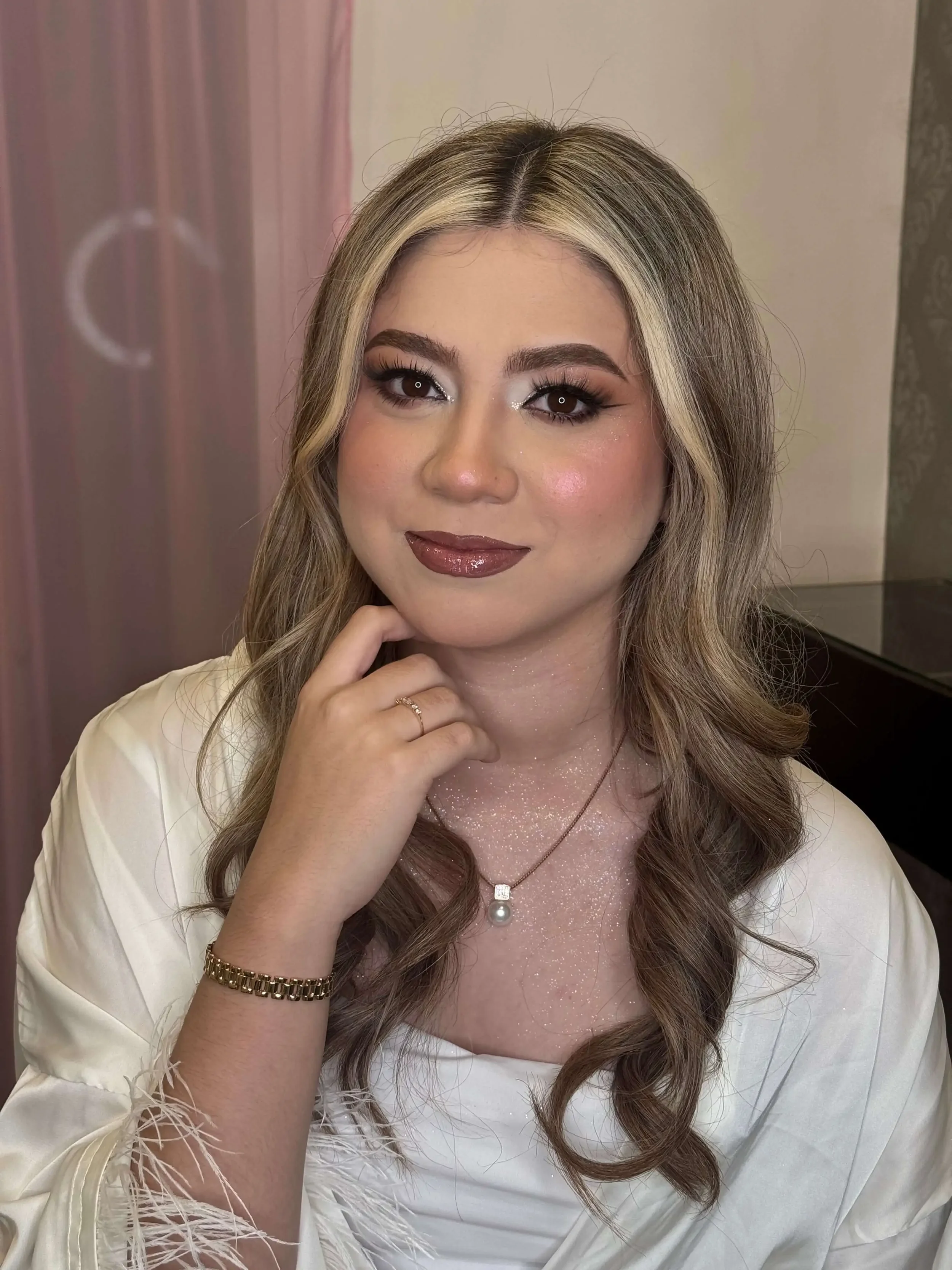 A portrait of a bride with a gentle smile, showcasing full-glam makeup with defined, smoky eyeshadow, fluttery lashes, and a glossy, dark rosewood-brown lip.
