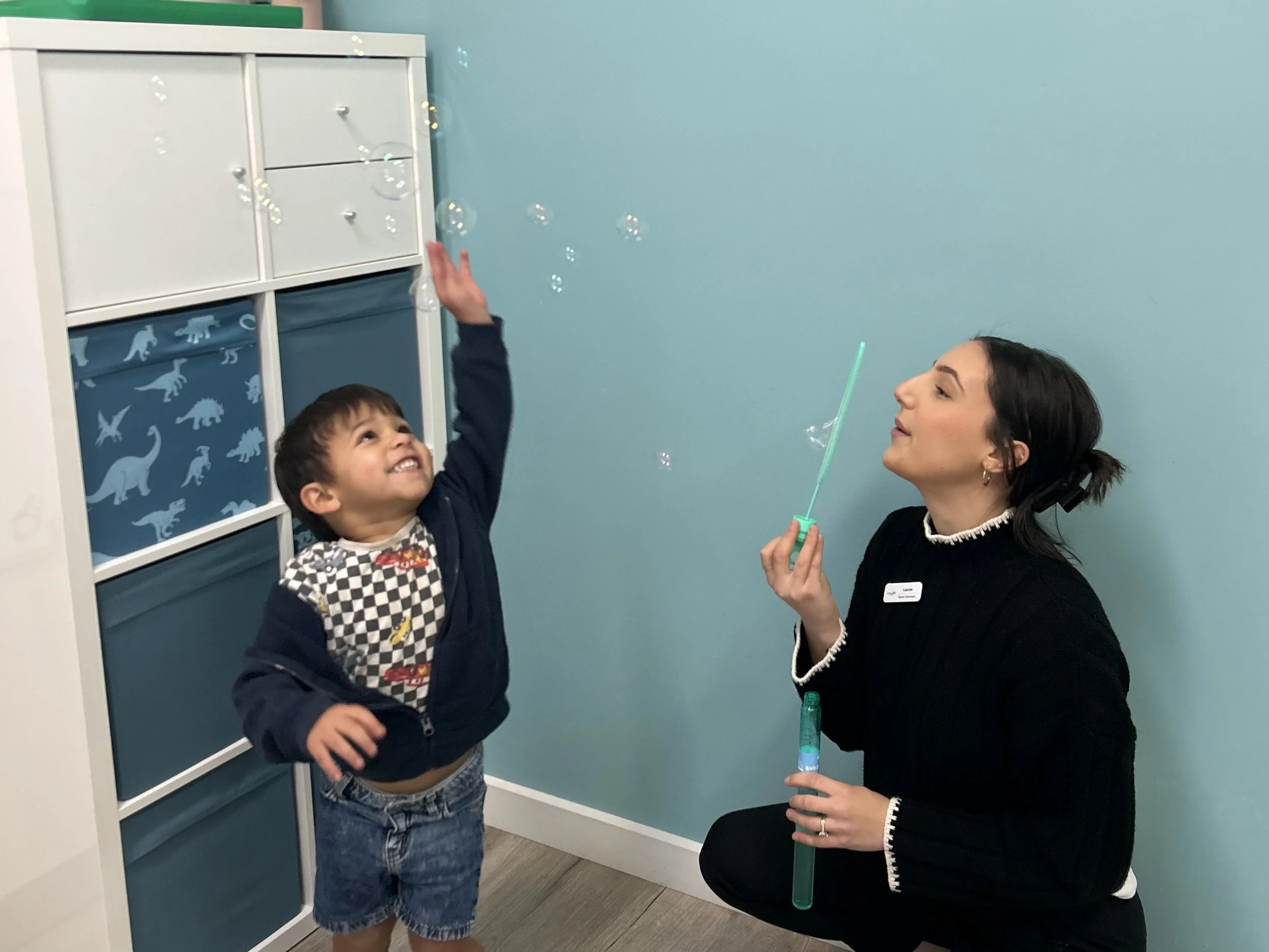 A young boy and a woman blowing bubbles in a room with a blue wall and a white storage unit.