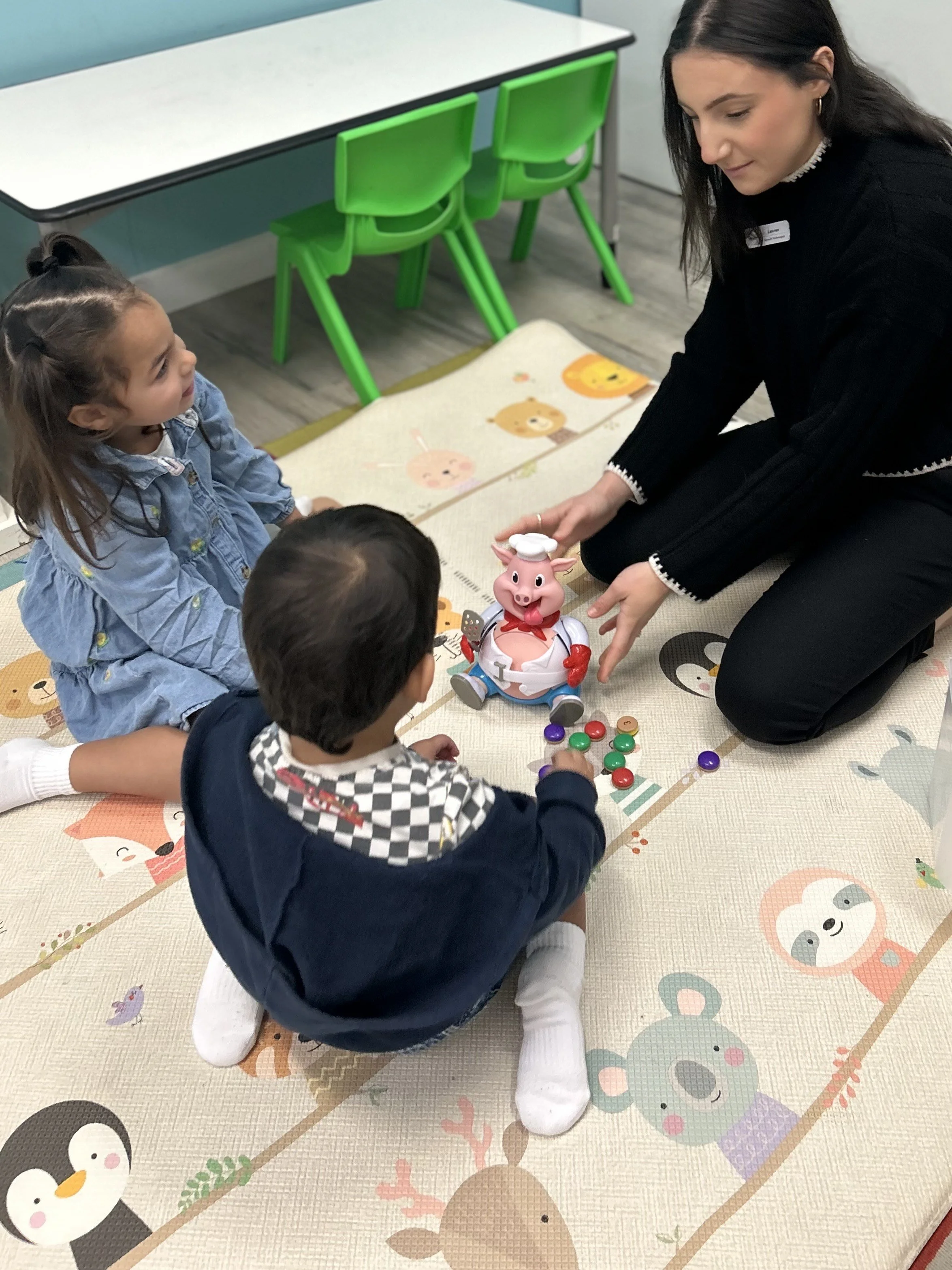A teacher and three young children playing a game with a pig toy robot on a colorful animal-themed carpet in a classroom.