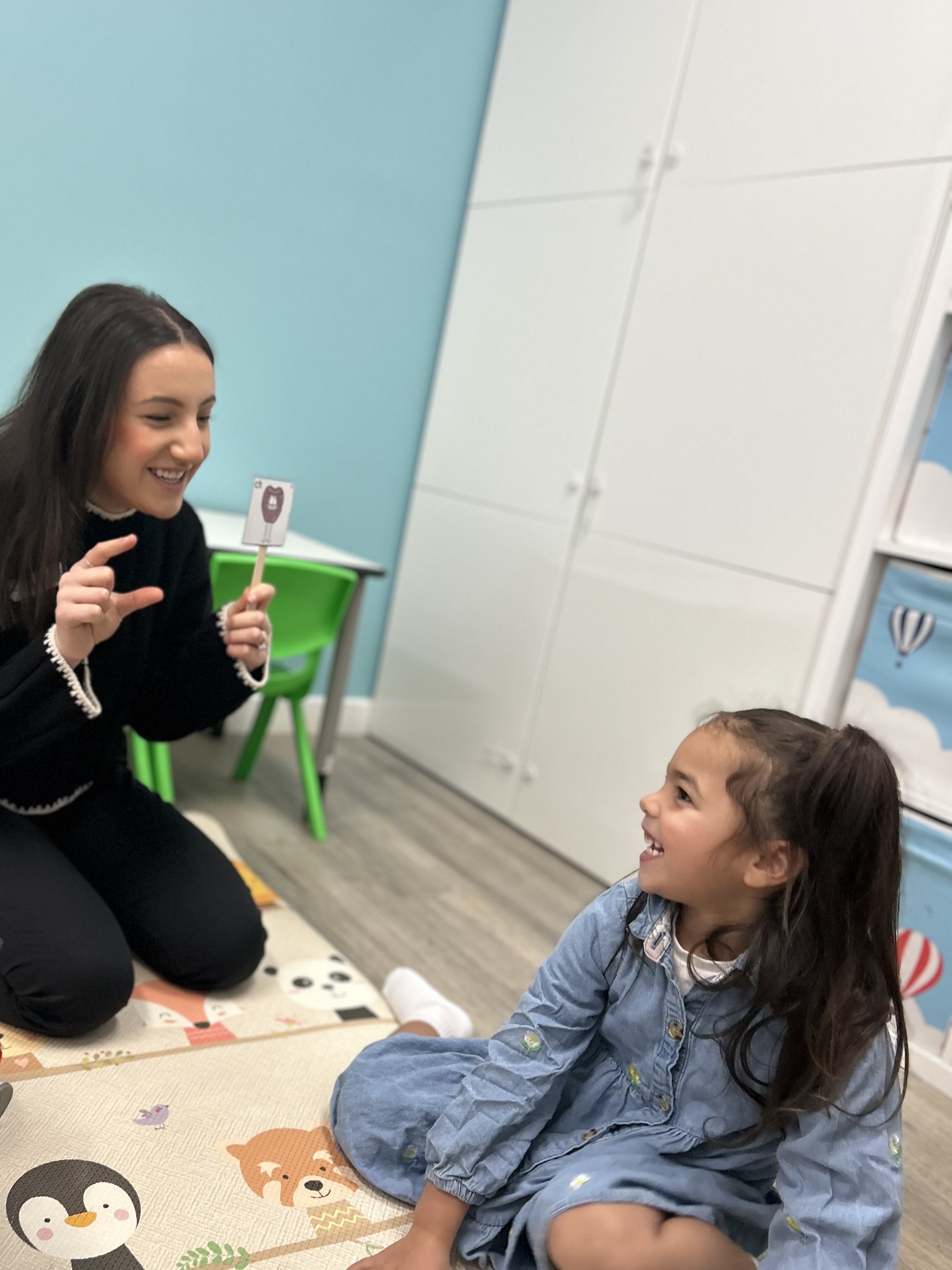 A woman with long dark hair kneeling and a young girl with long curly hair sitting on a colorful mat, both smiling and looking at each other. The woman holds a card with a cartoon bear illustration.