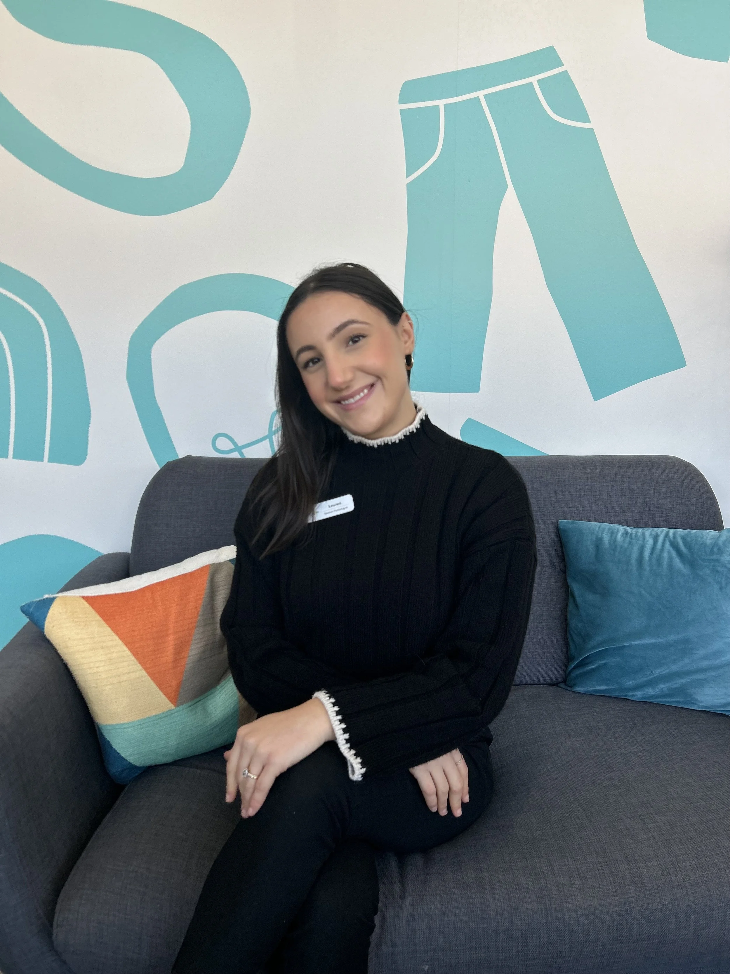 A woman sitting on a gray sofa, smiling, with a colorful pillow and a blue pillow, against a background wall with blue abstract clothing shapes.
