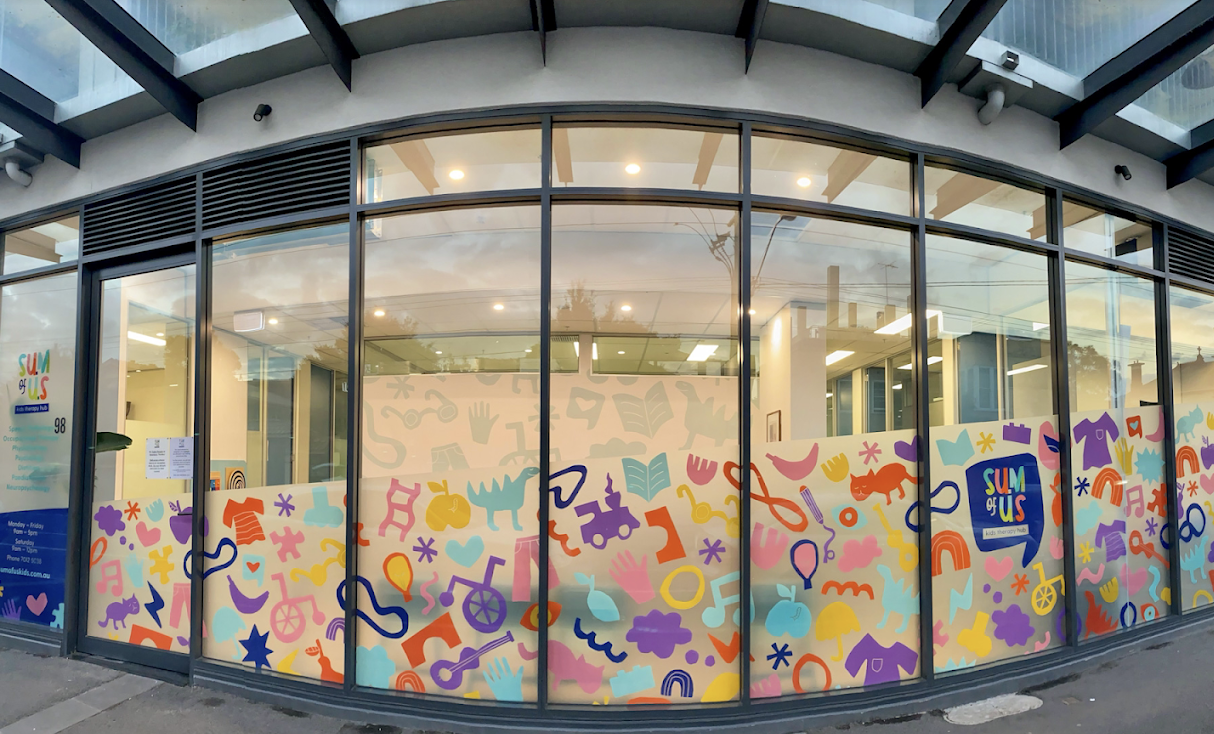 Exterior view of a building with large glass windows featuring colorful abstract patterns of animals, hearts, stars, clouds, and musical notes, with a logo that reads "Sun GUS."