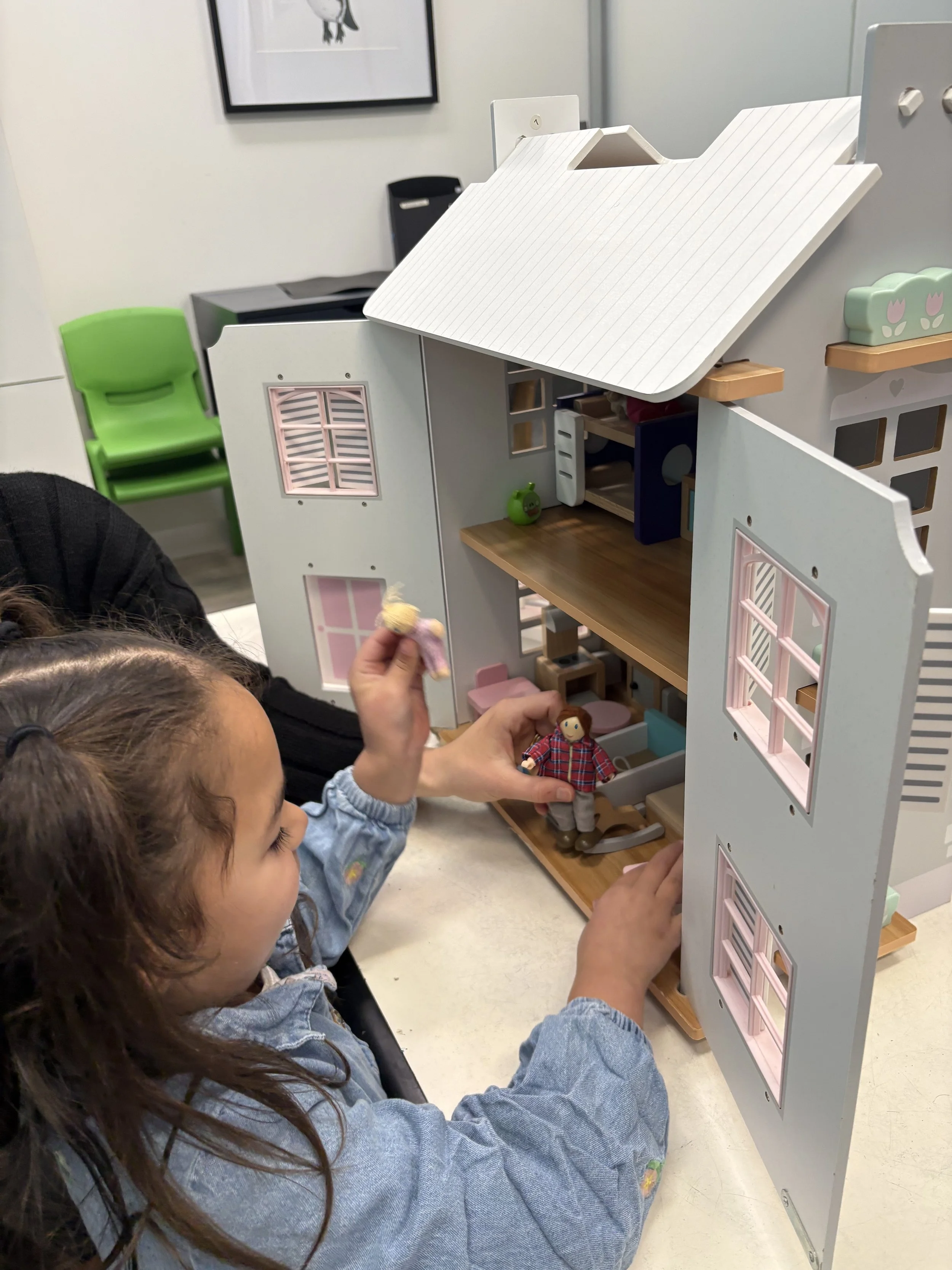 Children playing with a dollhouse indoors, one child holding a doll and another with a doll on the table.