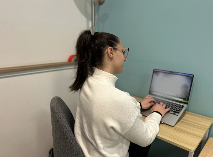 A woman with glasses and a white sweater working on a laptop at a wooden desk in a room with white and teal walls.