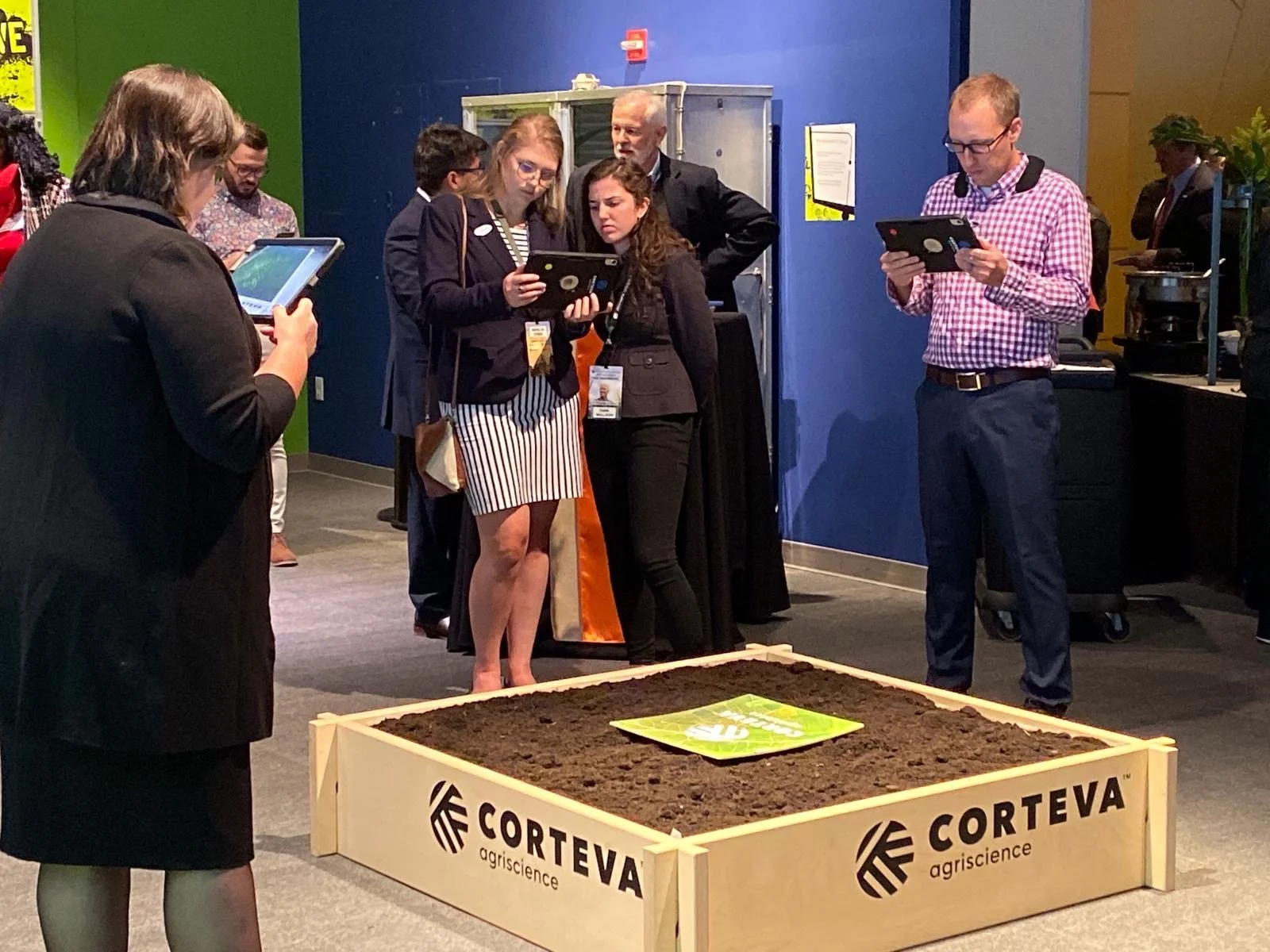 Group of people looking at a large, soil-filled wooden box with Corteva Agriscience branding at an indoor event.