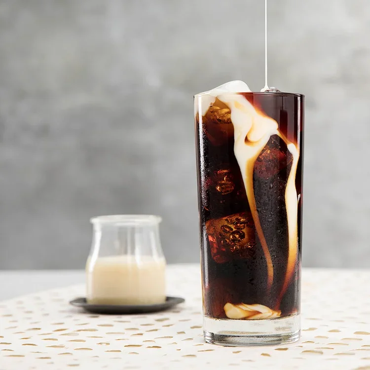 A glass of iced coffee with cream pouring into it, placed on a table with a small pitcher of cream in the background.