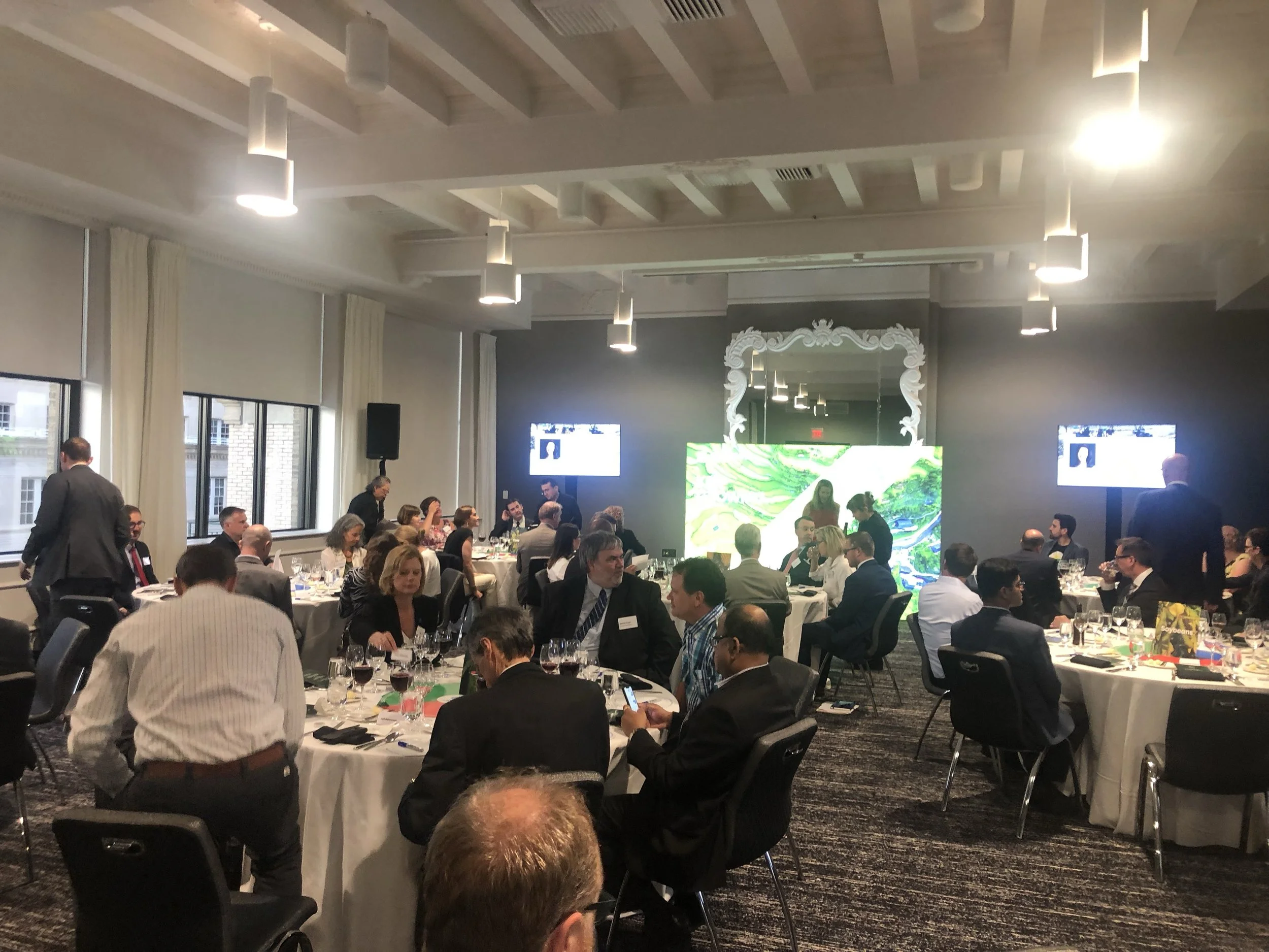 A crowded banquet hall with round tables, people dressed in formal business attire, large windows, and screening monitors on the walls.
