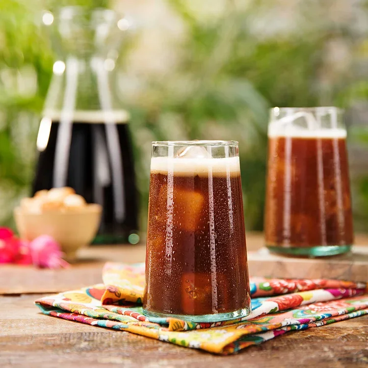Two glasses of iced dark soda with foam on top on a colorful cloth on a wooden table, with a blurred background of greenery and a coffee maker.
