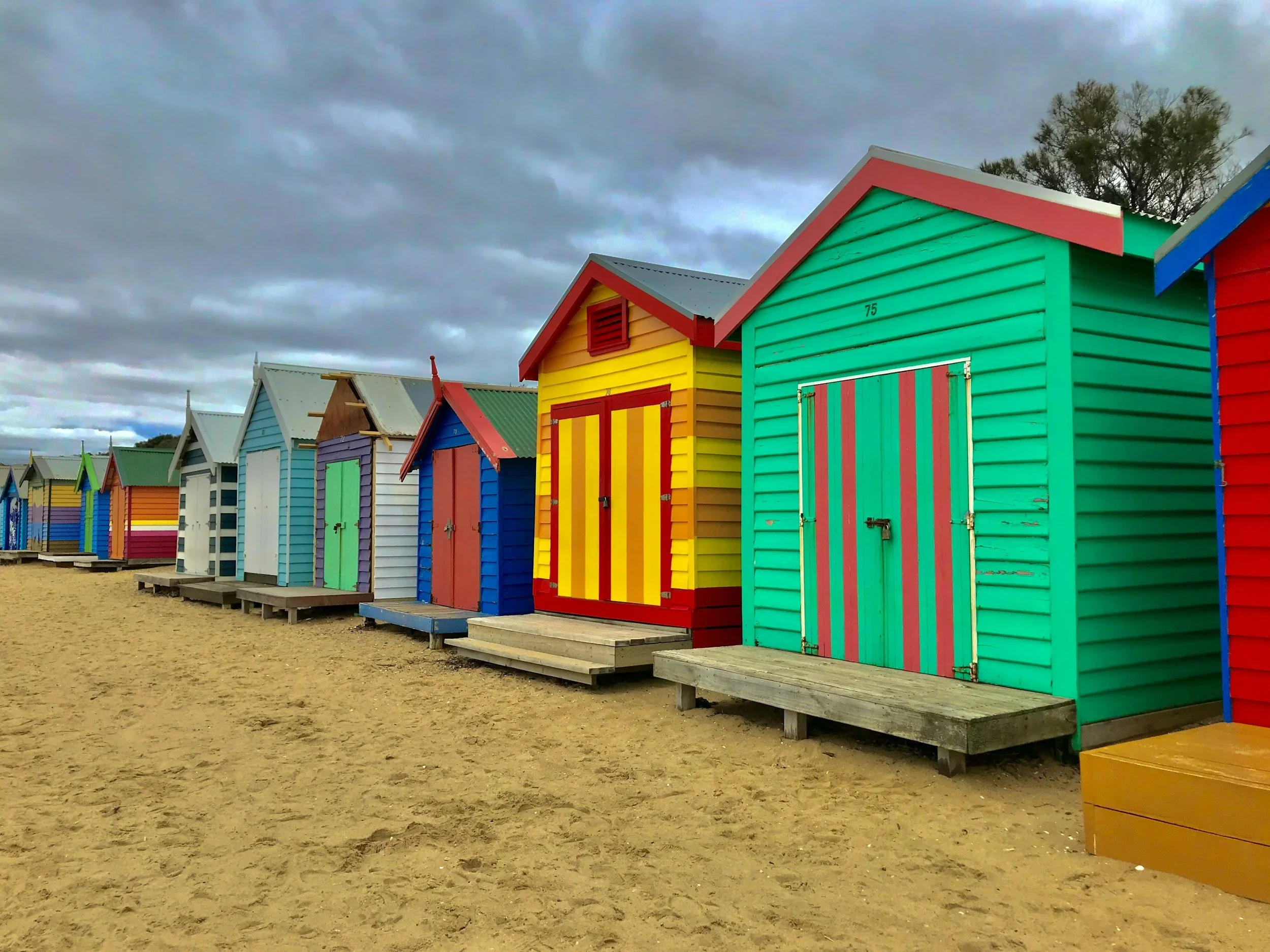 Colorful beach huts lined up on sandy beach under cloudy sky.