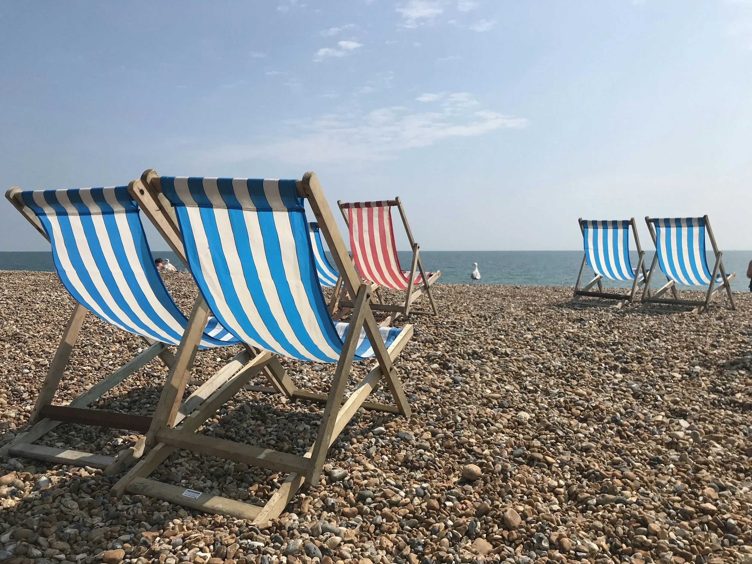 Beach scene with wooden deck chairs with blue and white striped fabric, and one red and white striped fabric, on a pebbled beach near the ocean under a partly cloudy sky.