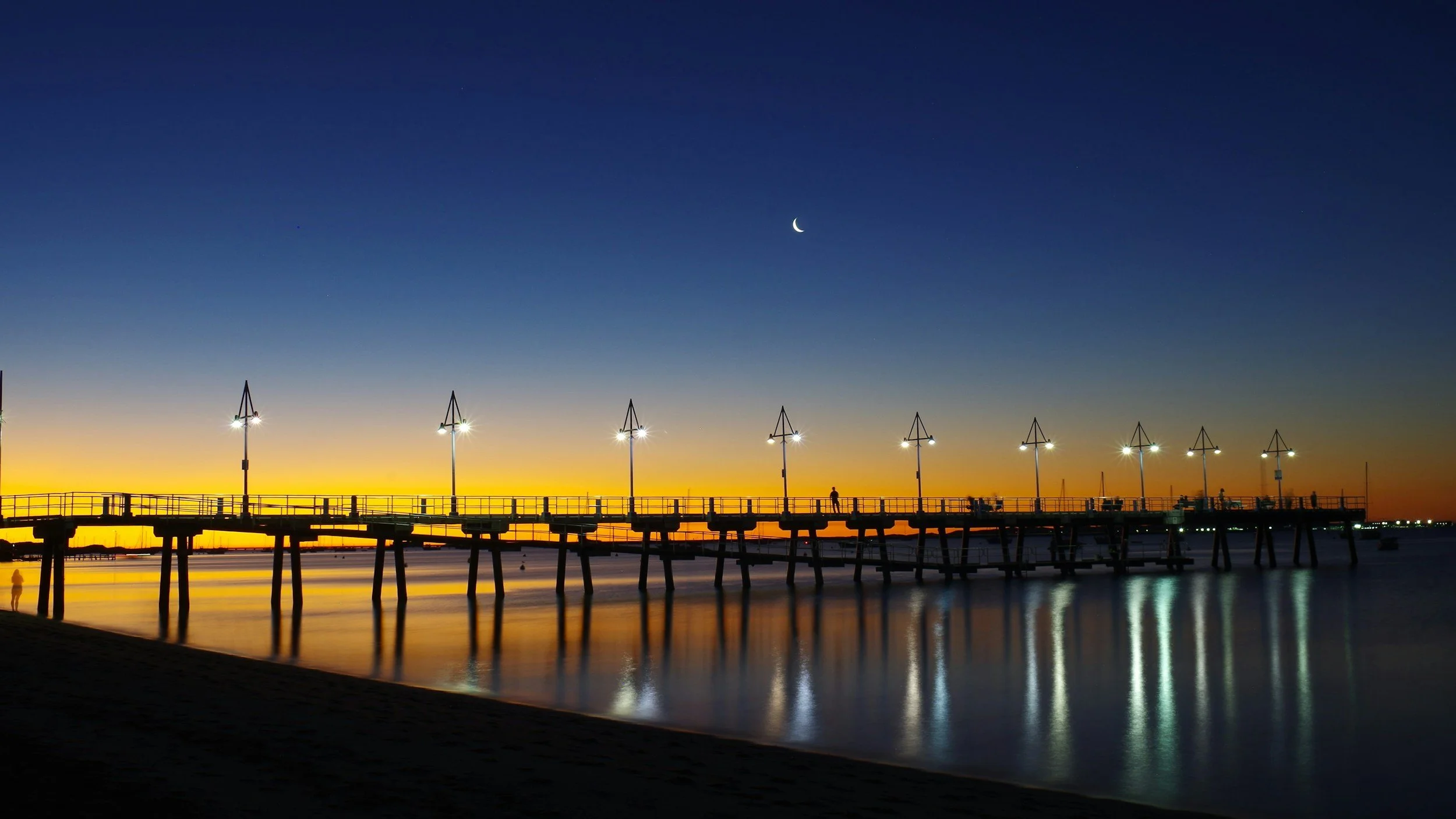 A pier extending over calm water at dusk, illuminated by streetlights with a crescent moon in the sky.