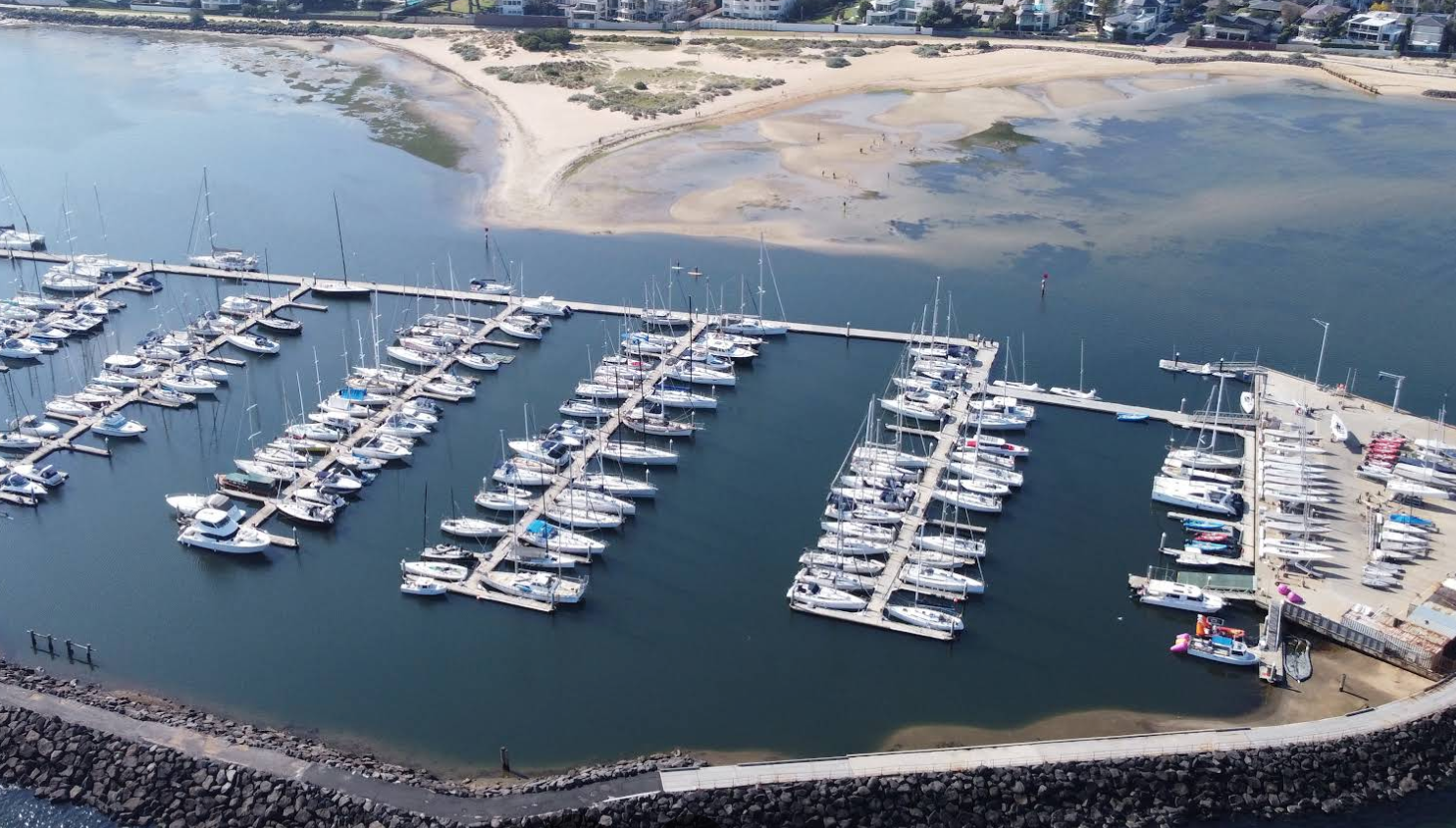 Aerial view of a marina with numerous sailboats and yachts docked in organized slips, adjacent to a sandy beach with a few people, and nearby residential buildings.