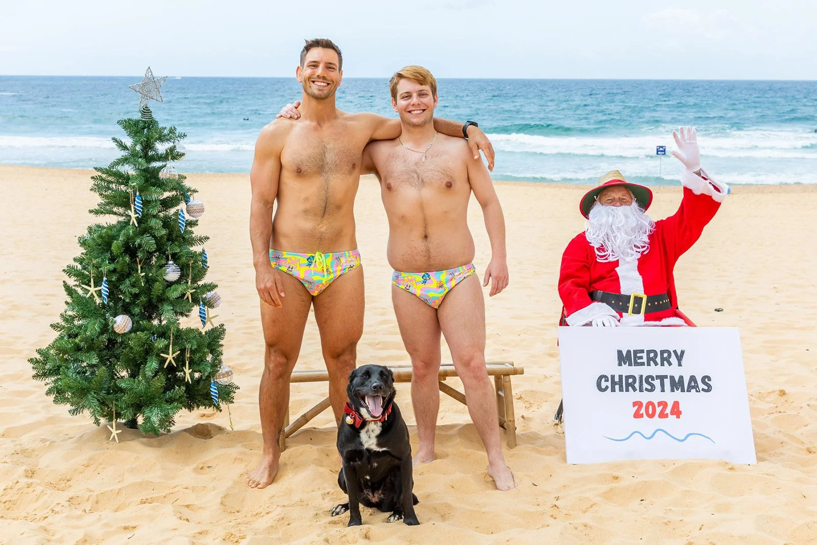 Two shirtless men wearing colorful swim briefs standing on a sandy beach with a black dog sitting in front of them. One man has his arm around the other. To the right, a person dressed as Santa Claus is sitting and waving, holding a sign that reads '