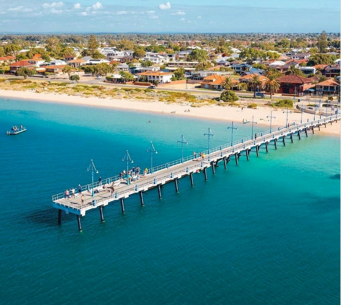 An aerial view of a long pier extending into calm turquoise waters near a sandy beach and a residential area with houses and palm trees.