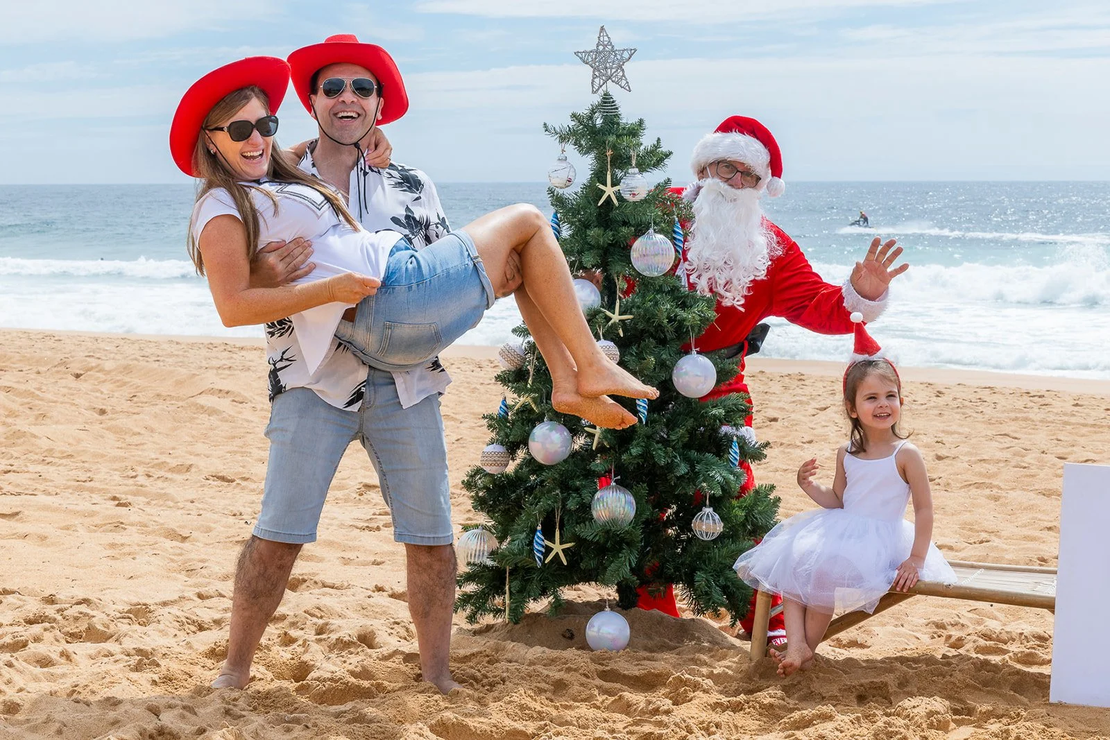 A family dressed in beach attire celebrating Christmas on the beach, with a decorated Christmas tree and Santa Claus.