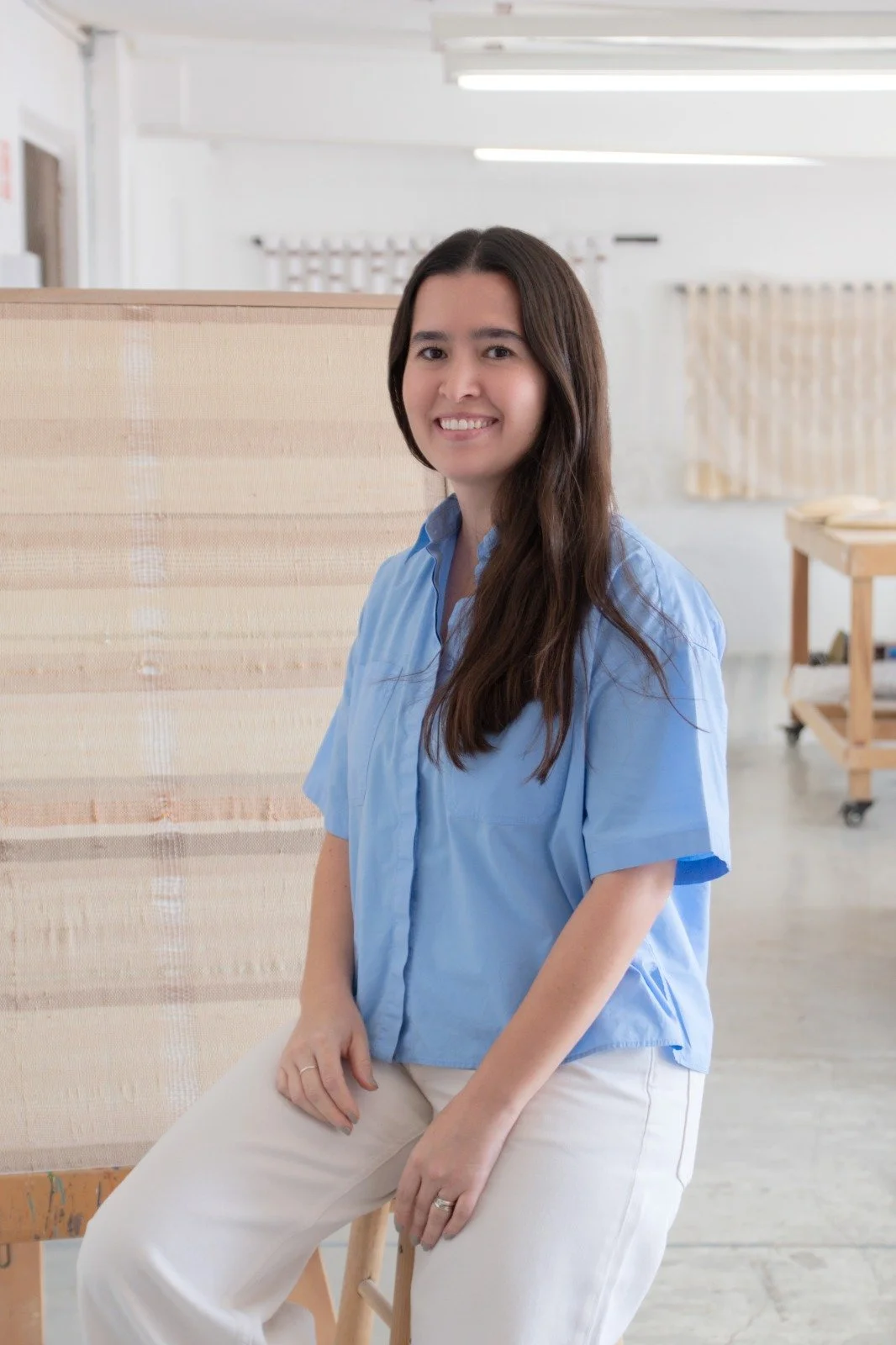 Mujer con cabello largo y castaño, sonriendo, usando blusa azul, en un espacio interior que parece una oficina o taller, con mobiliario de madera y cortinas claras.