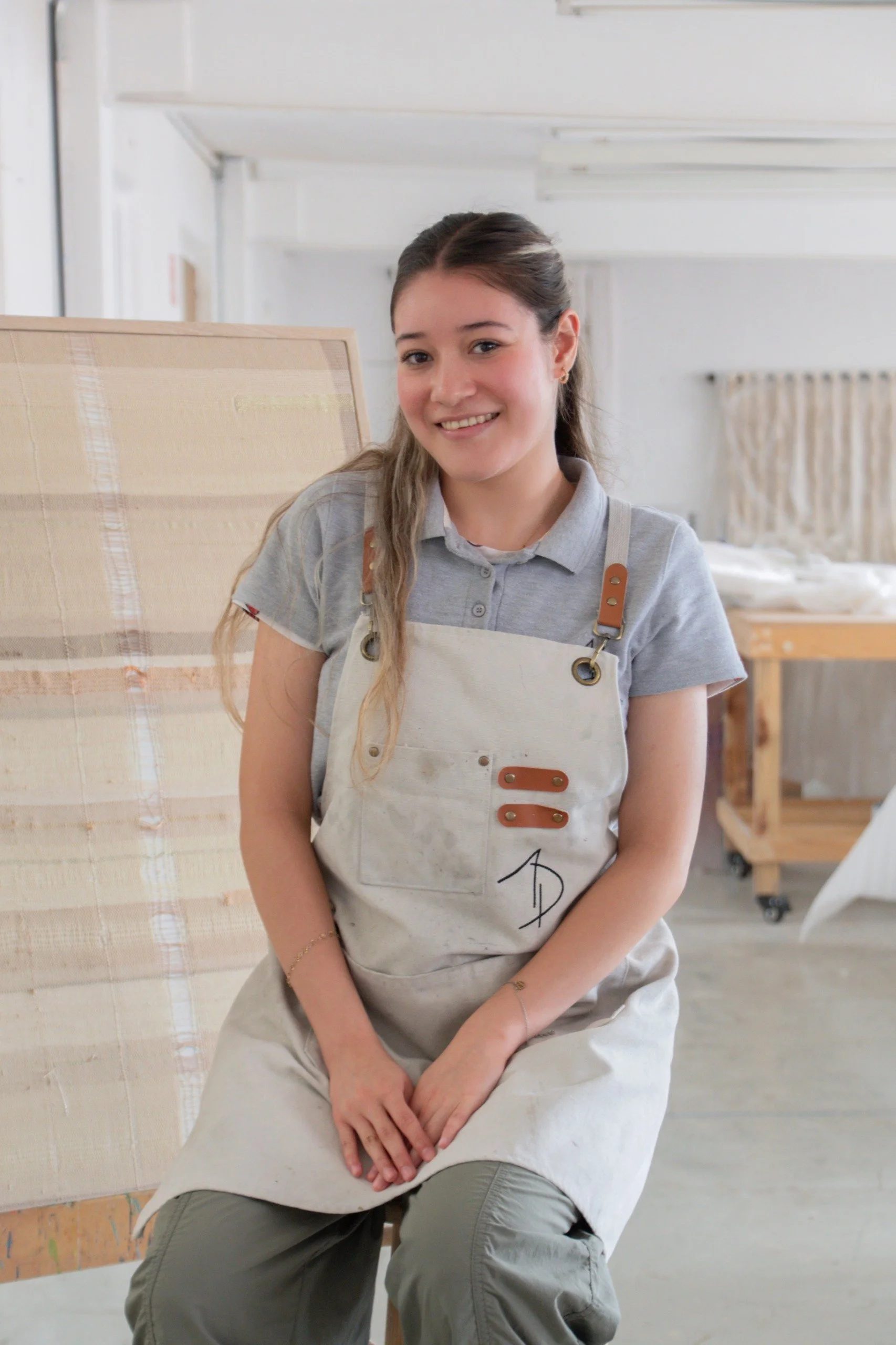 Una joven mujer con cabello castaño claro y coleta, sonriendo, lleva una camiseta gris y un delantal beige, en un espacio de taller con mobiliario de madera y tela de lienzo en el fondo.