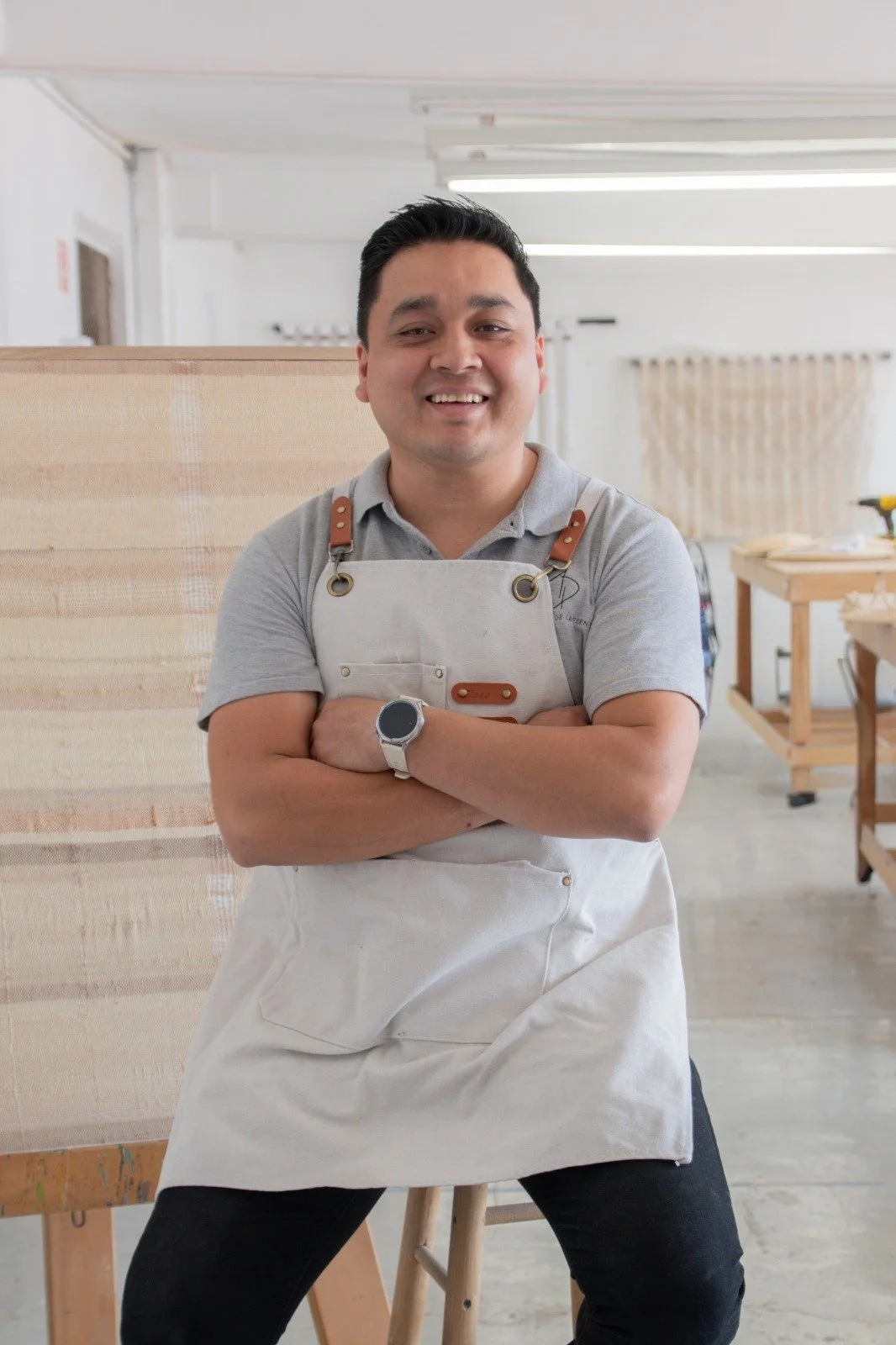 Hombre sonriendo con brazos cruzados, usando delantal en taller de carpintería.