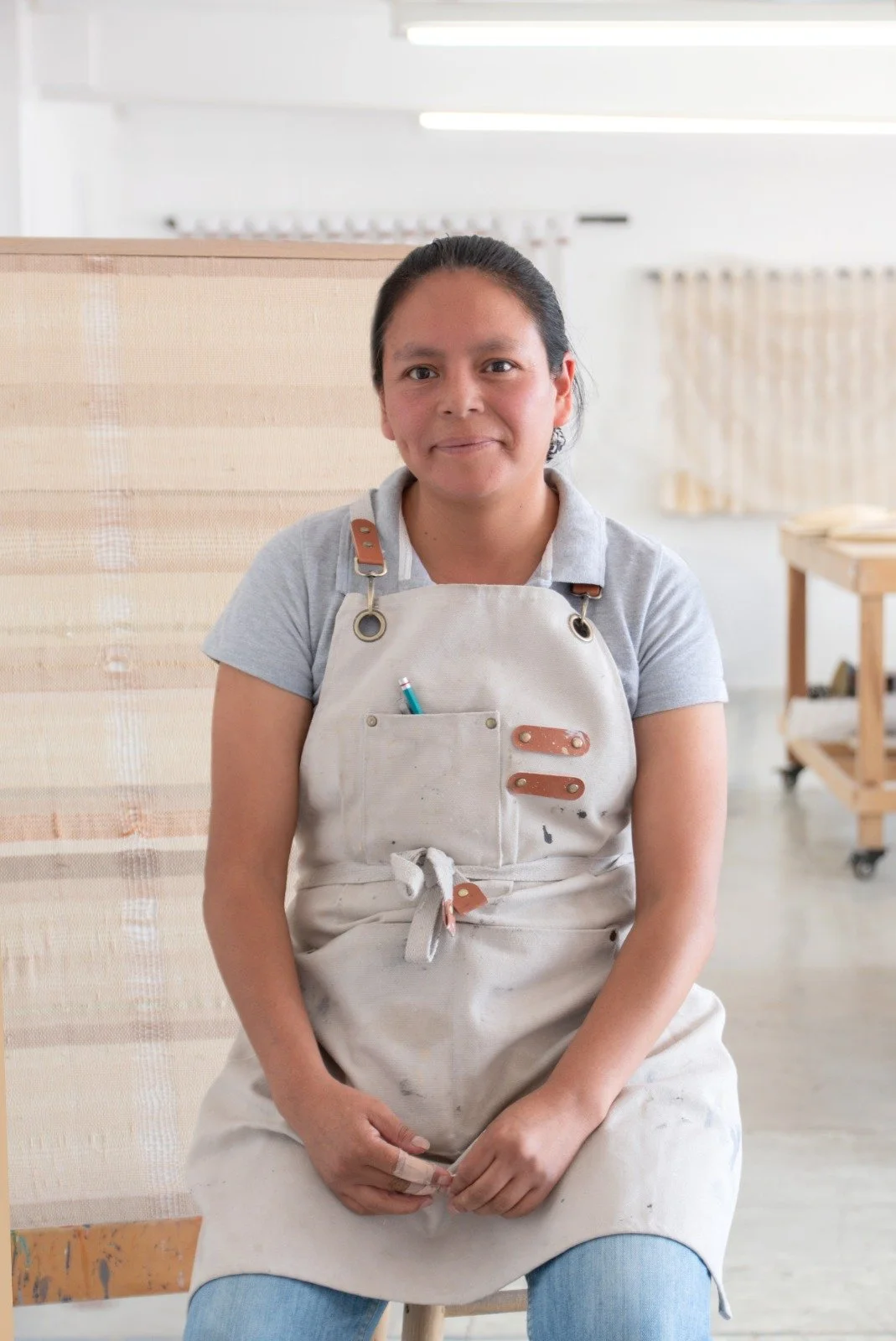 Mujer sentada en un taller de carpintería o artesanía, lleva delantal, cabello recogido y sonríe.