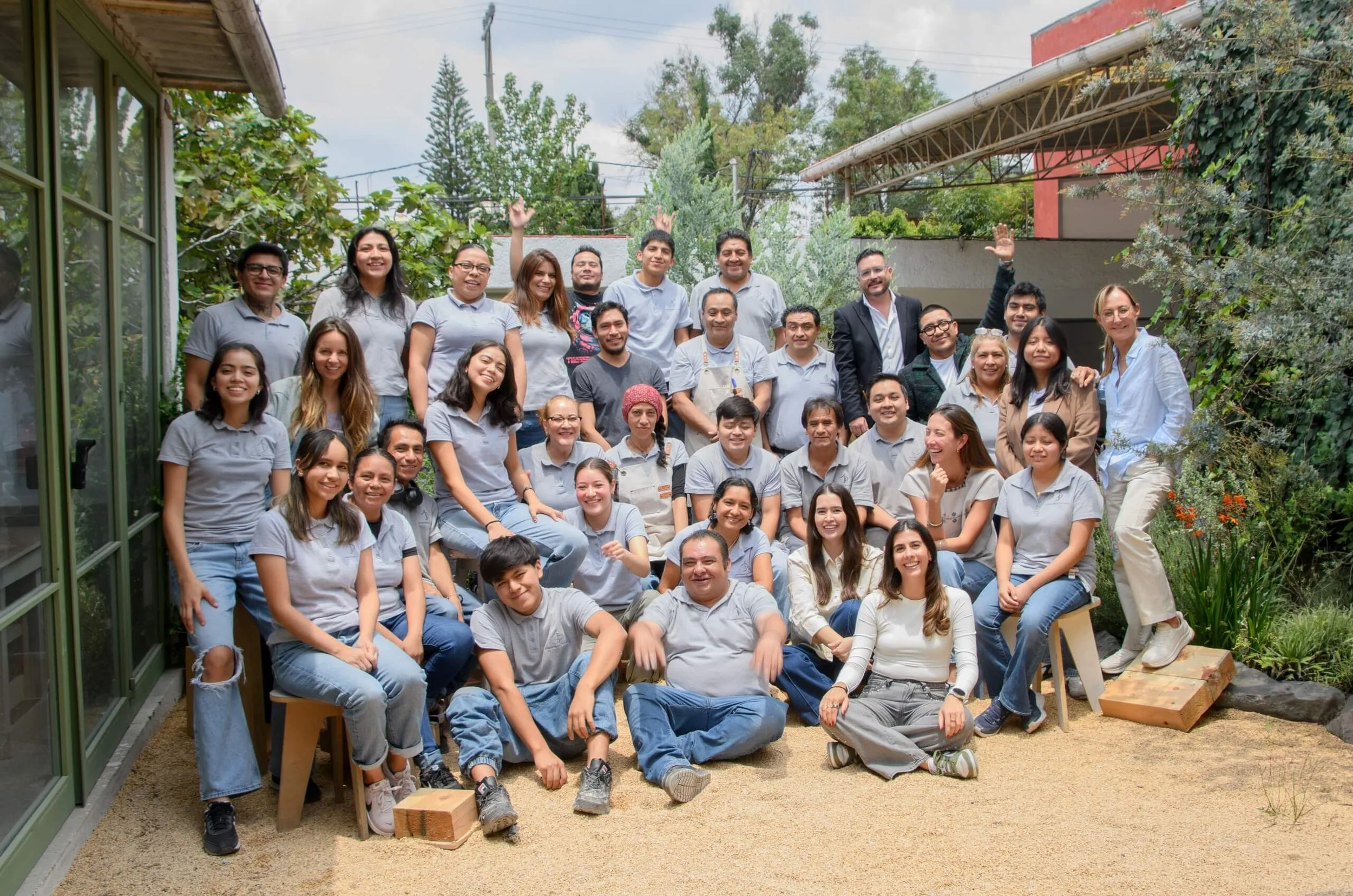 Grupo de personas posando y sonriendo en un jardín, algunos están sentados y otros de pie, con árboles y plantas de fondo.