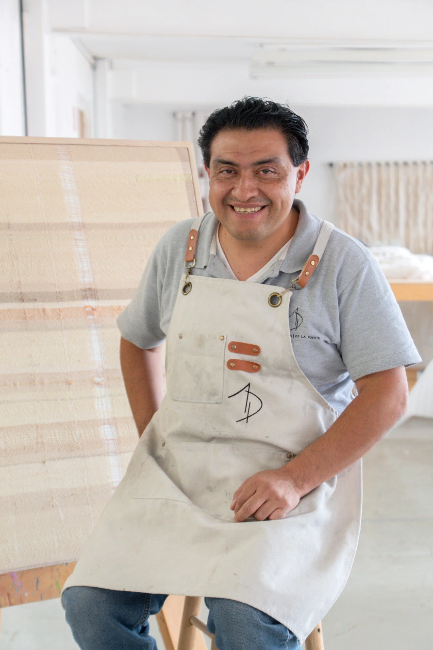 Hombre sentado en una silla, usando un delantal y sonriendo, en un ambiente de estudio de arte o pintura.