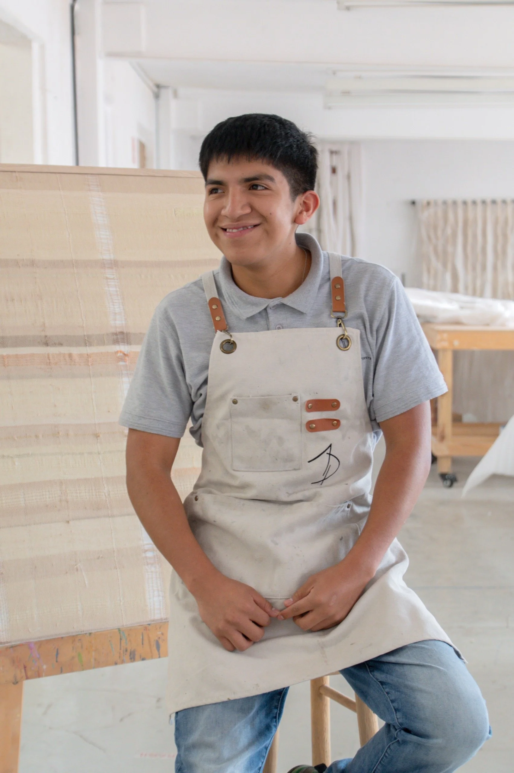 Joven con camiseta gris y delantal claro, sentado frente a un caballete en un estudio de arte, sosteniendo su delantal con ambas manos, con una sonrisa en el rostro.