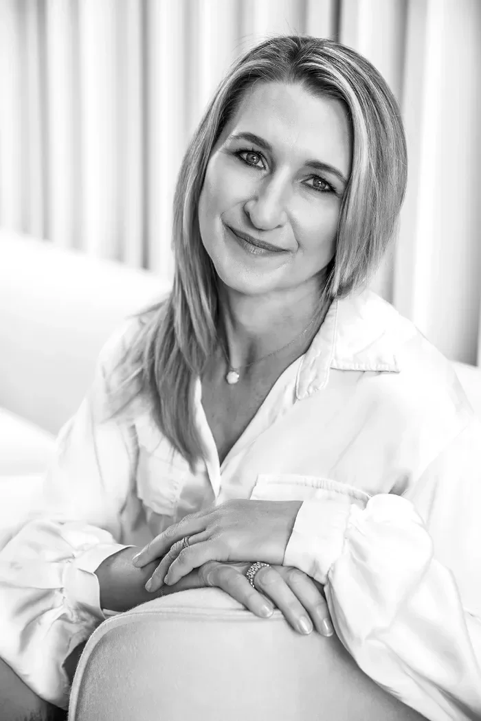 A woman with shoulder-length hair smiling, sitting with her hands resting on her lap, wearing a white shirt, in a black-and-white portrait.