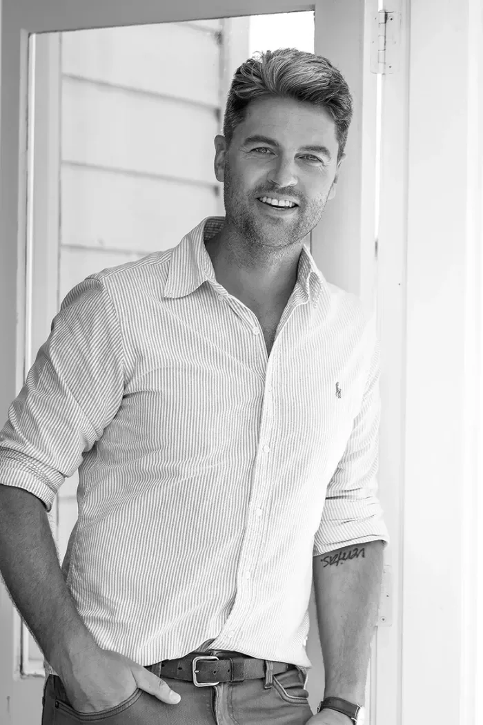 Black and white photo of a smiling man with short hair, wearing a light striped button-up shirt with rolled-up sleeves, standing outside near a house, with his hand in his pocket.