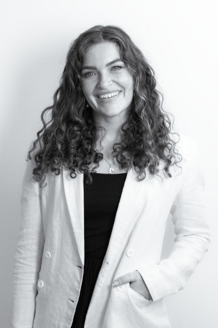 A young woman with long, curly hair smiling and winking, wearing a white blazer over a black top, and a necklace with a cross pendant.